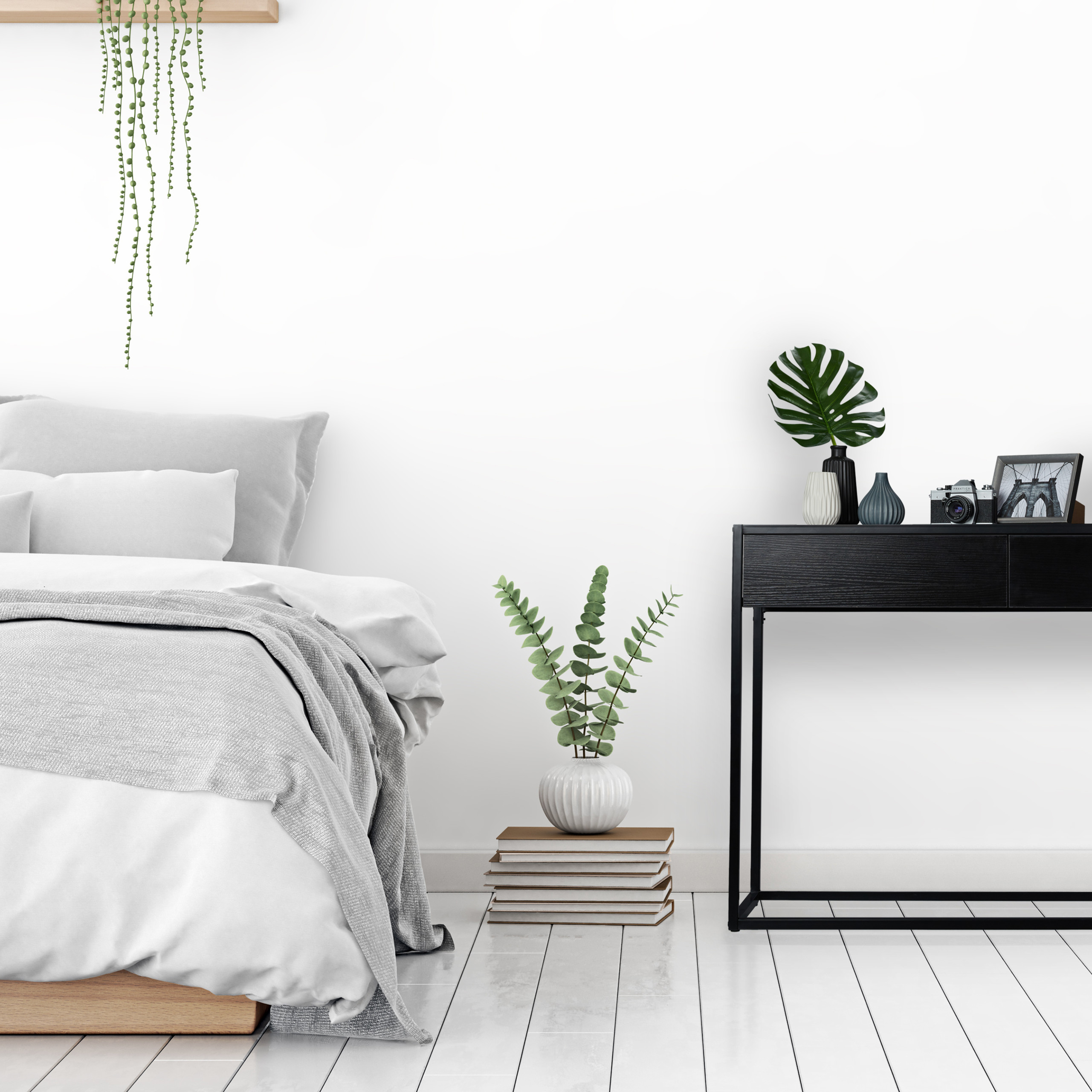 Ein minimalistisches Schlafzimmer mit Bett, hängender Pflanze, Büchern und schwarzem Tisch gegen weiße Wand.