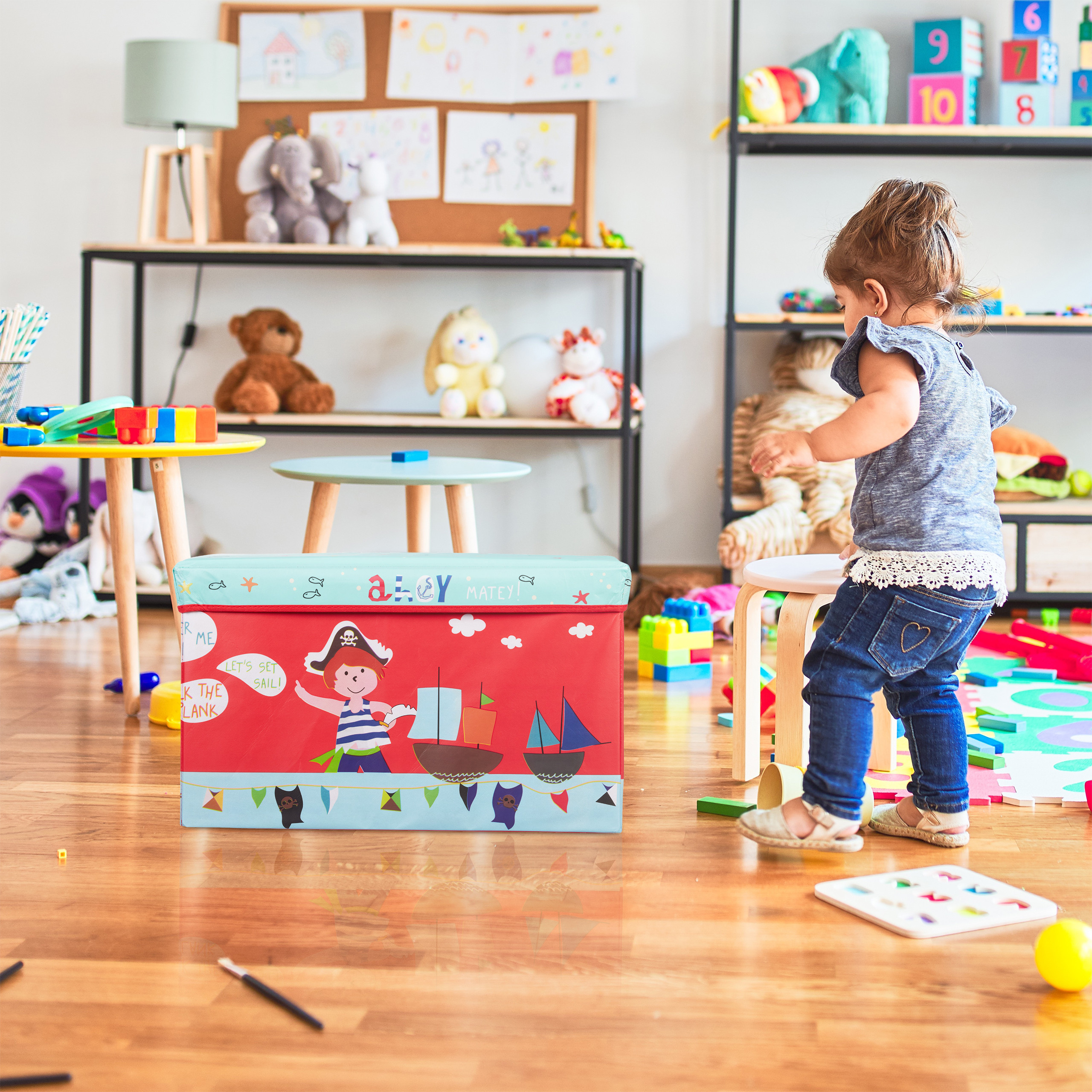 Kleinkind in blauem Oberteil spielt im hellen, mit Spielzeug gefüllten Kinderzimmer.