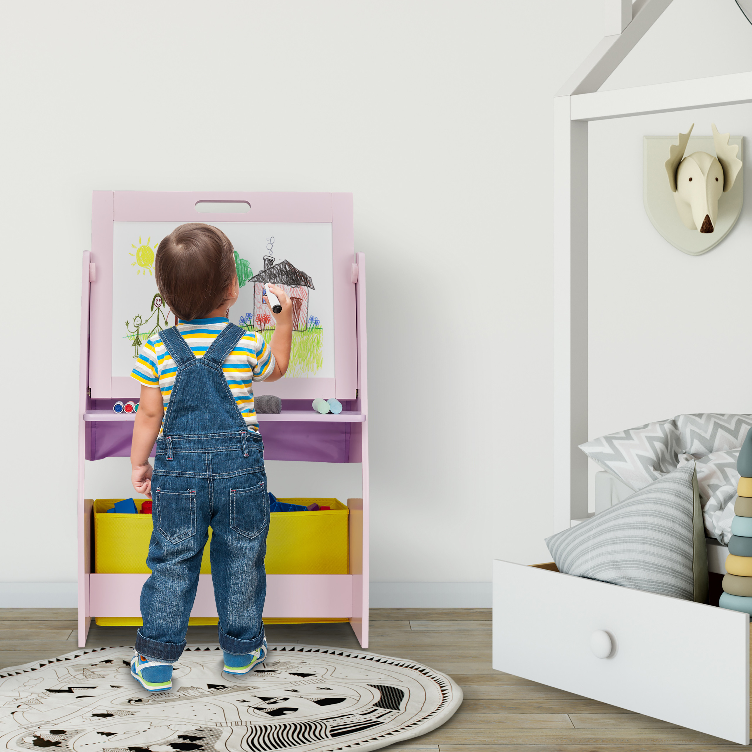 Ein Kleinkind in Jeanslatzhose malt auf eine rosa Staffelei in einem hellen, modernen Kinderzimmer.