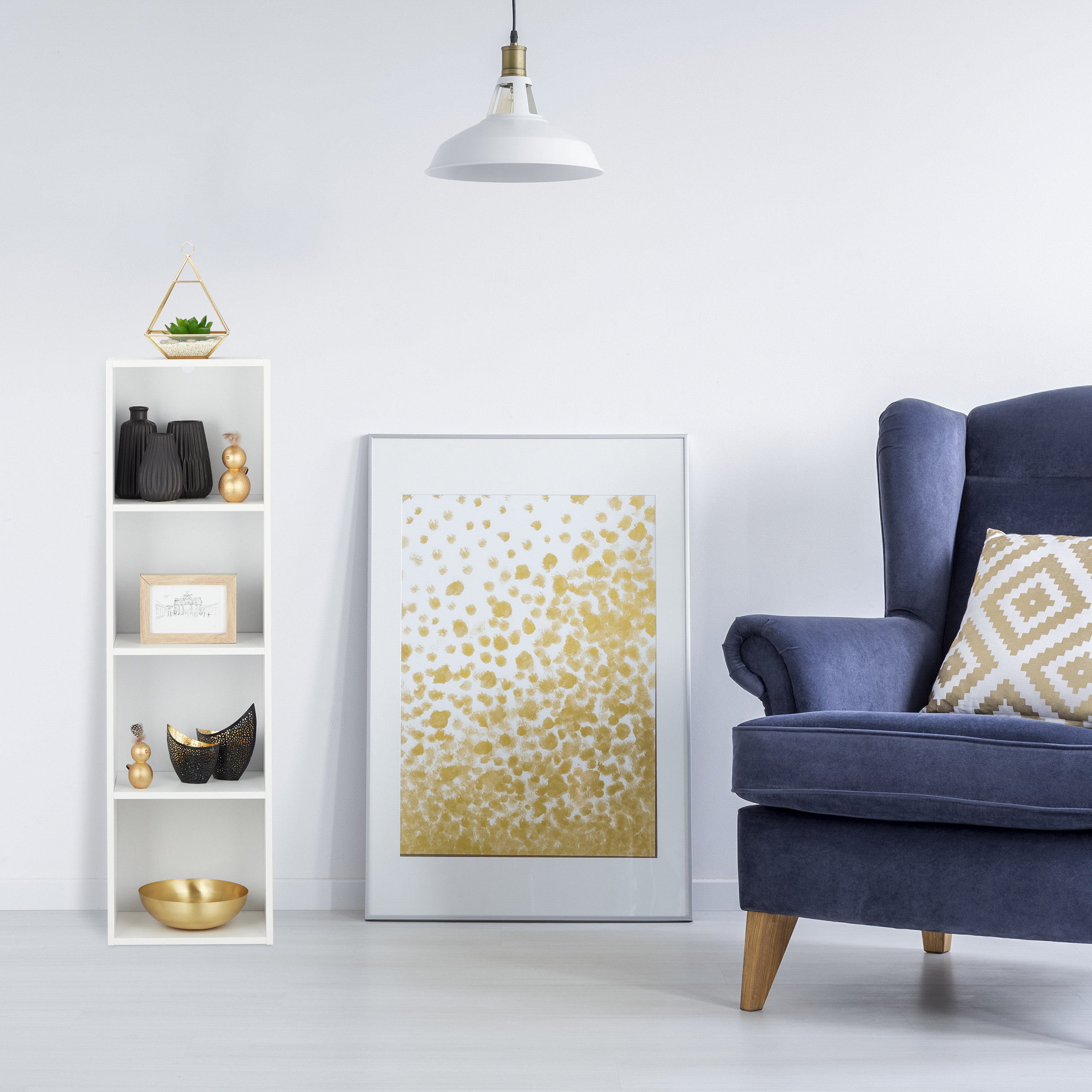 Ein blauer Sessel, ein weißes Regal mit Deko, ein großes goldenes gepunktetes Bild und eine weiße Lampe.