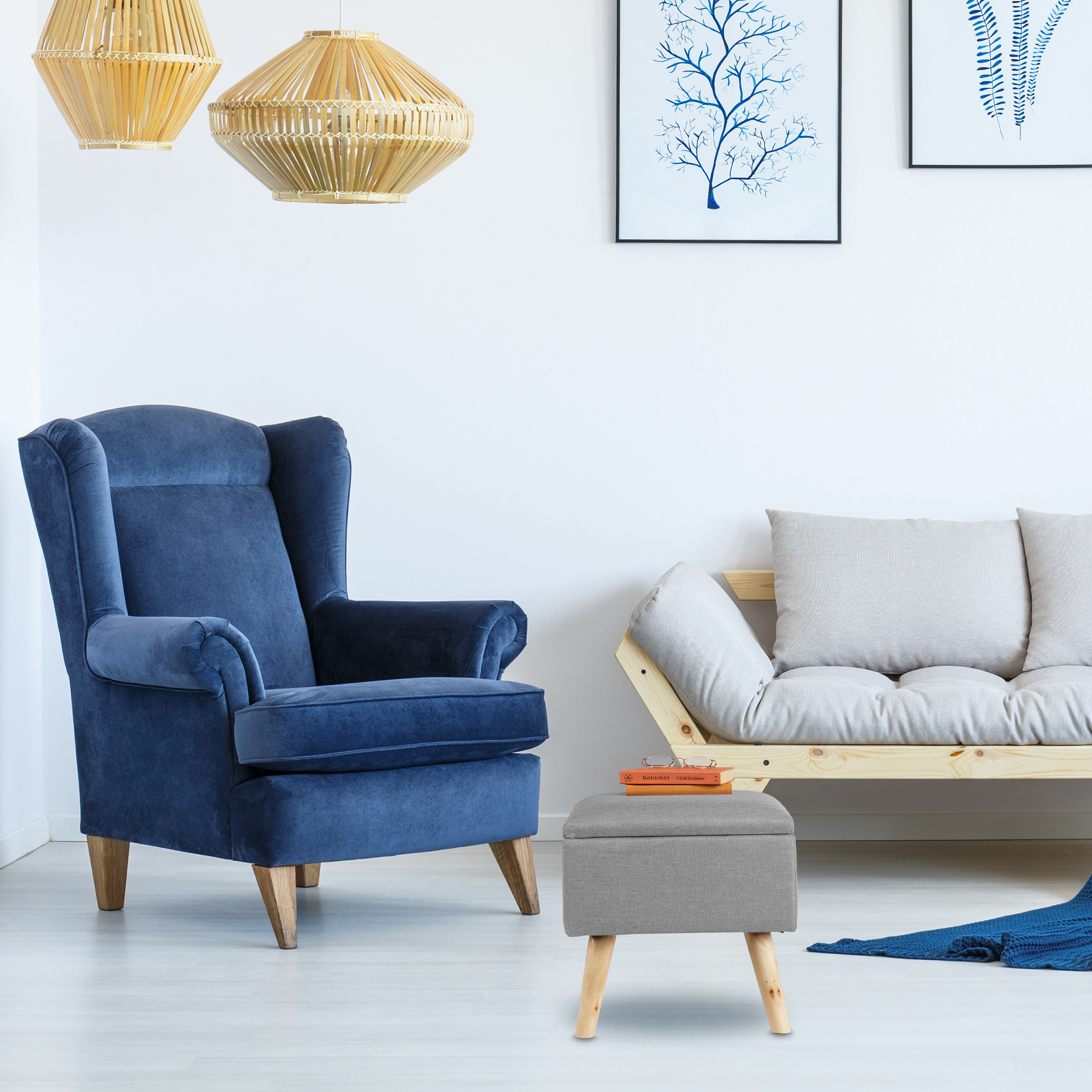 Ein blauer Sessel, ein heller Holzsofa, eine graue Fußbank mit Büchern und Wandkunst in einem weißen Raum.