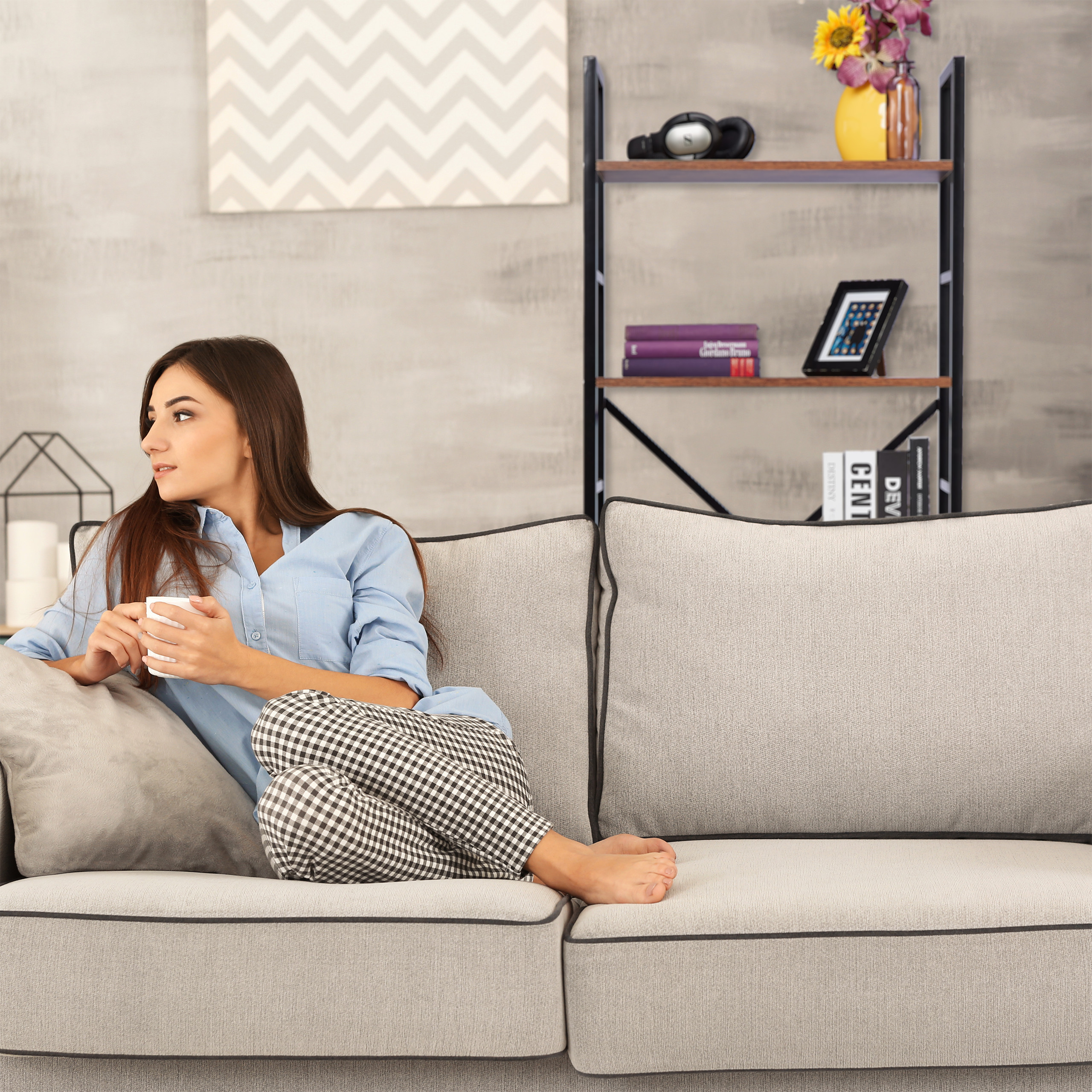 Eine Frau in blauem Hemd und karierten Hosen sitzt mit einer Tasse auf einem grauen Sofa vor einem Regal.