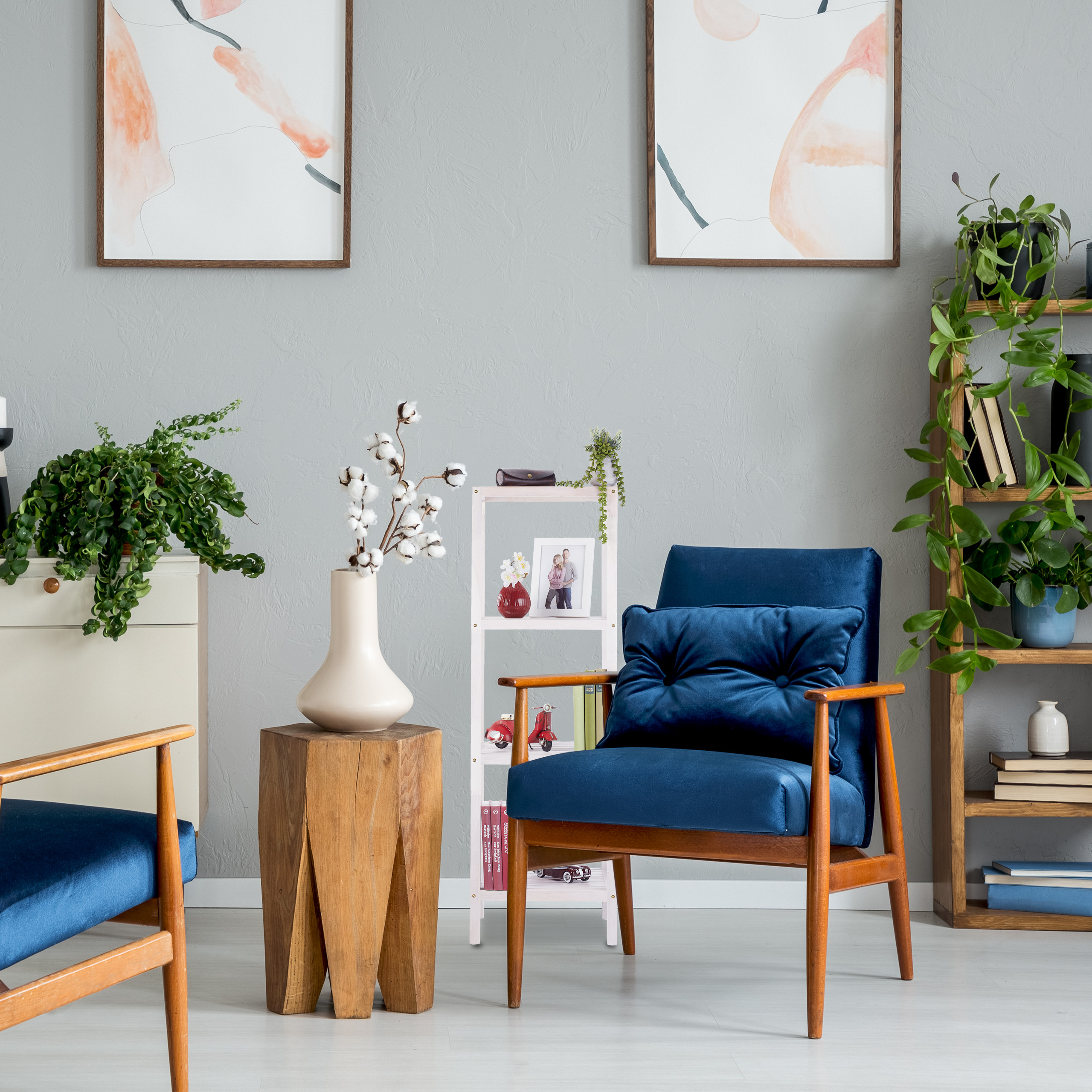 Wohnzimmer mit blauem Sessel, Holzhocker, Pflanzen und zwei abstrakten Bildern an der grauen Wand.