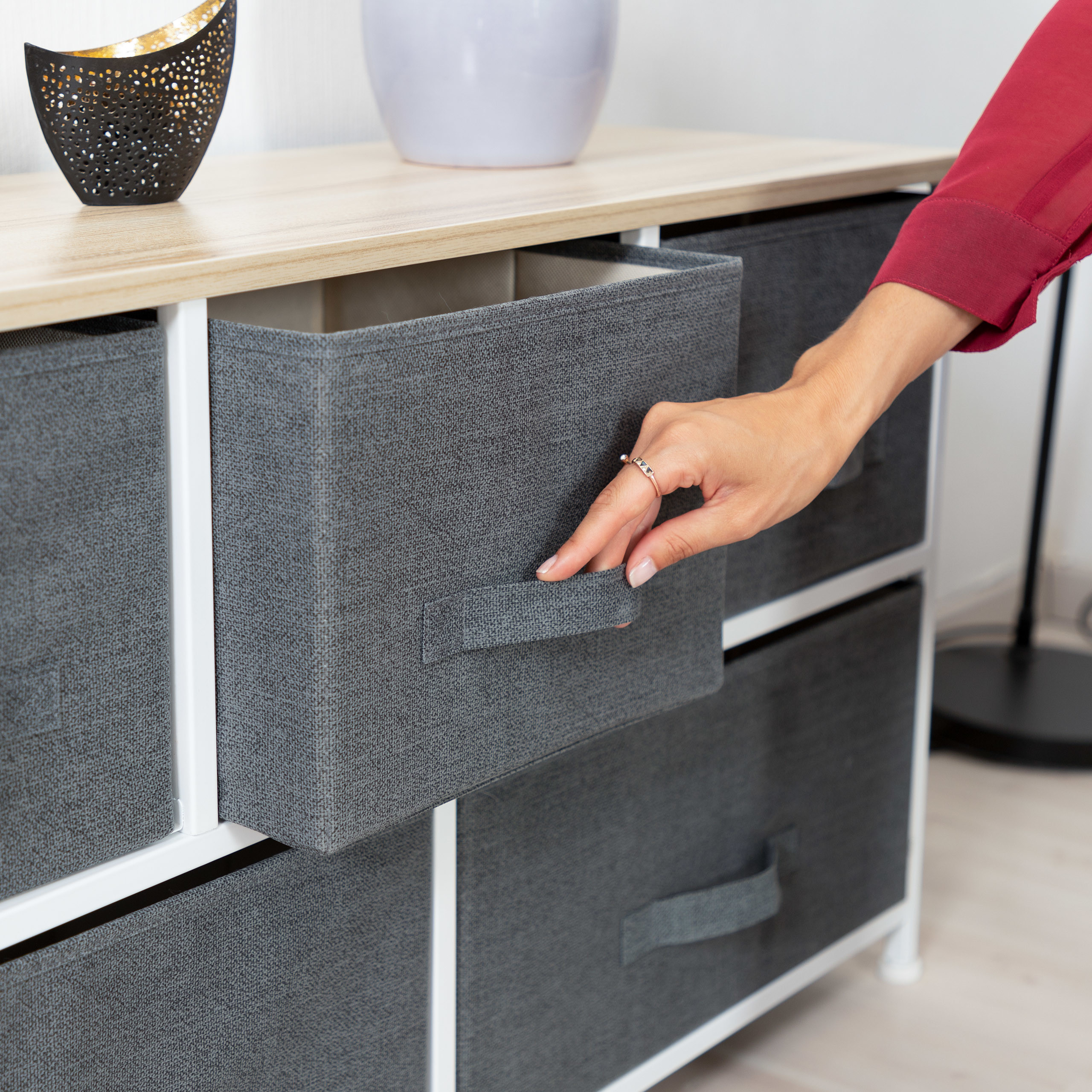 Eine Hand zieht eine graue Stoffschublade aus einer hellen Holzkommode mit weißem Metallgestell.