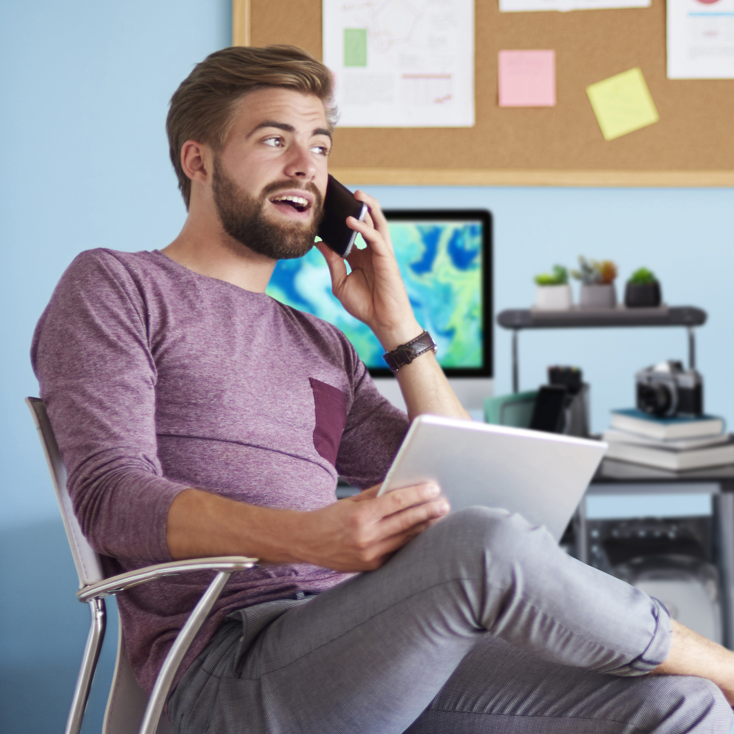 Ein bärtiger Mann im lila Pullover telefoniert und hält ein Tablet im Büro.