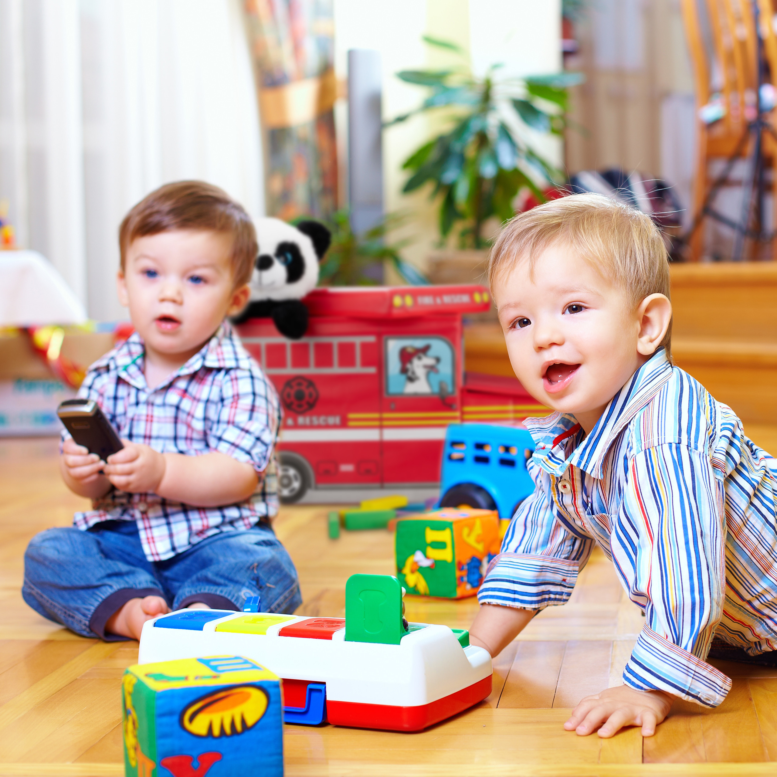 Zwei Kleinkinder spielen auf einem Holzboden mit Spielzeug, während im Hintergrund ein Feuerwehrauto steht.