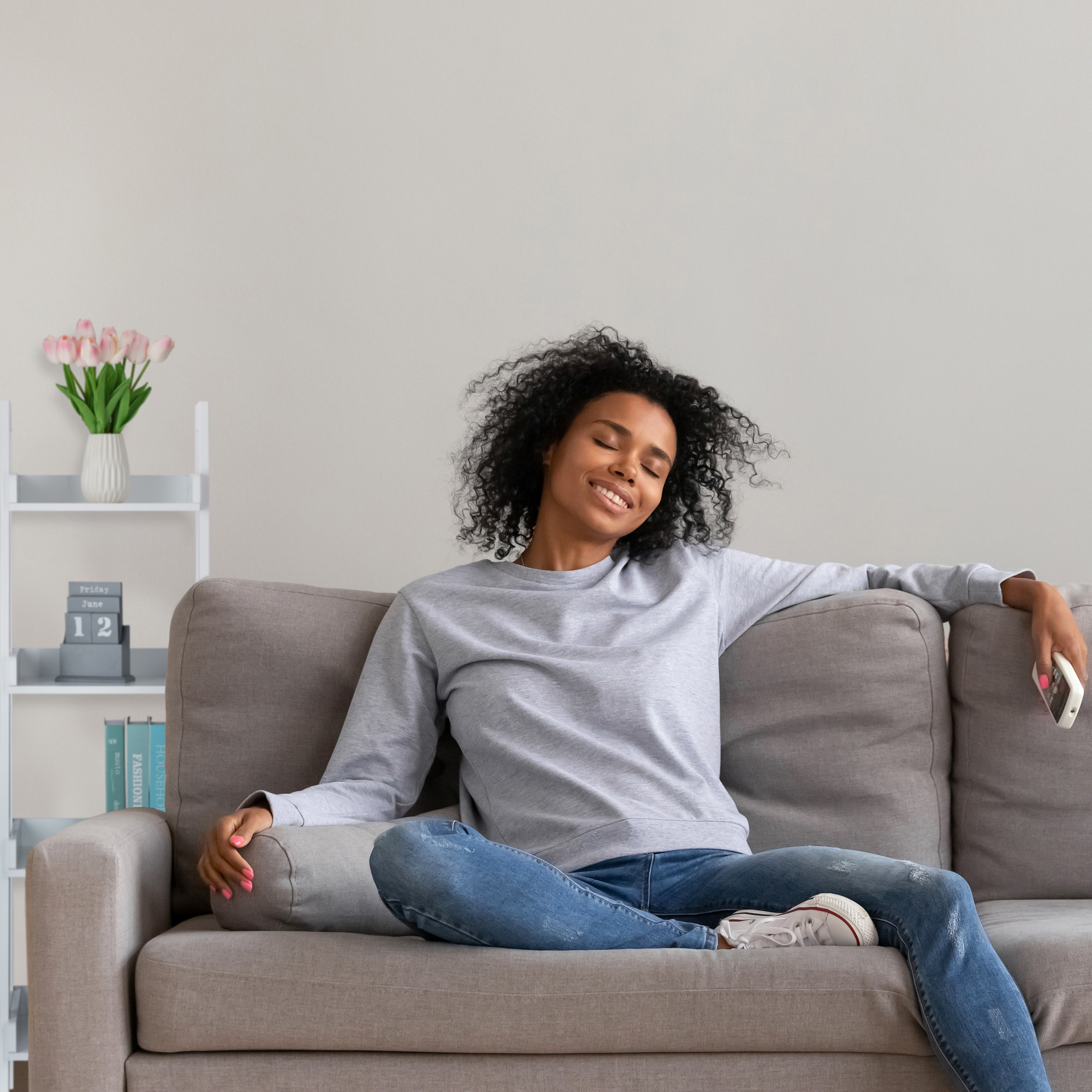 Eine Frau sitzt entspannt auf einem grauen Sofa mit einem Smartphone in der Hand.