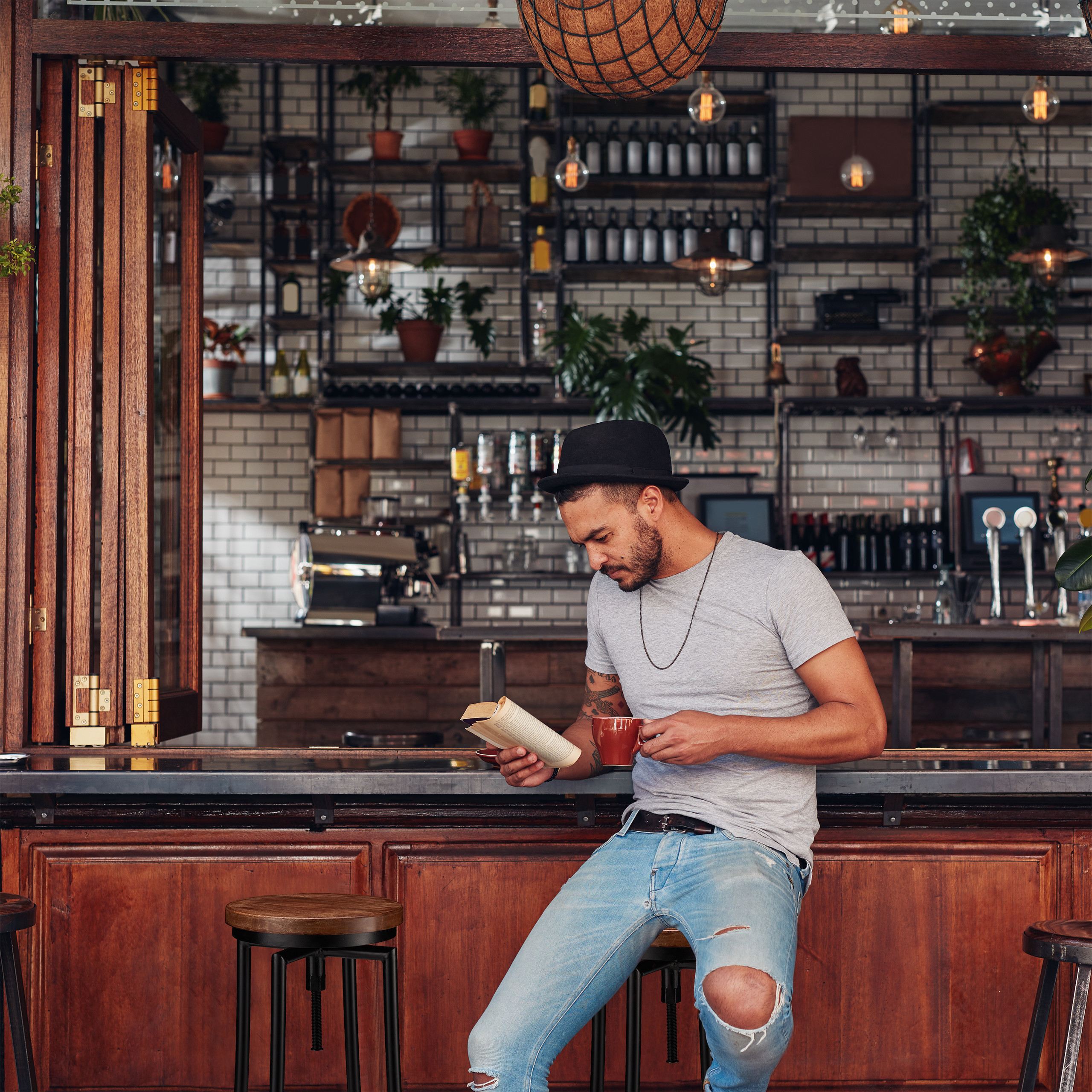 Ein Mann mit Hut und grauem Shirt liest ein Buch und hält eine Tasse an einer Bar.