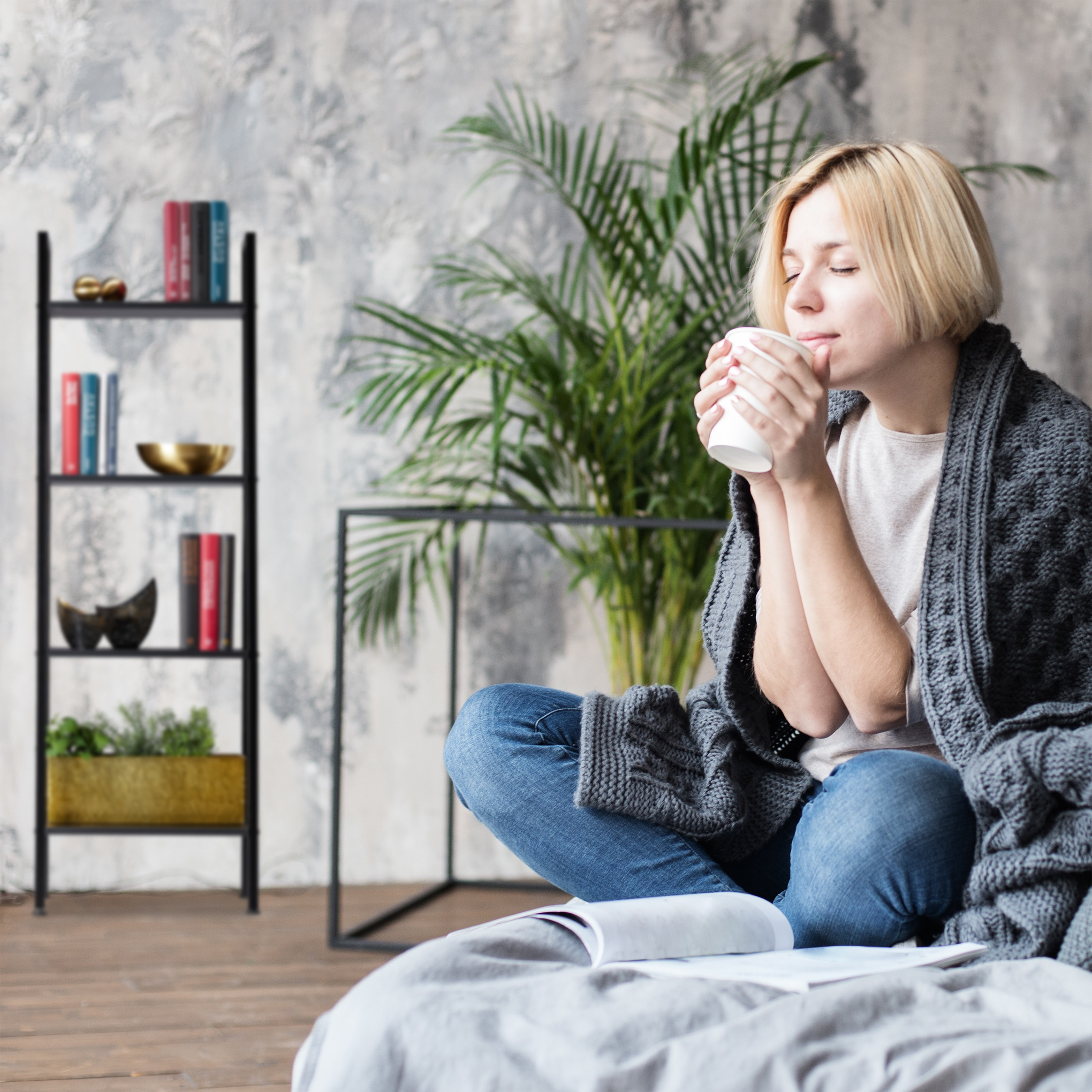 Eine blonde Frau in einer grauen Decke sitzt im Schneidersitz, hält eine weiße Tasse und riecht daran neben einer Pflanze.