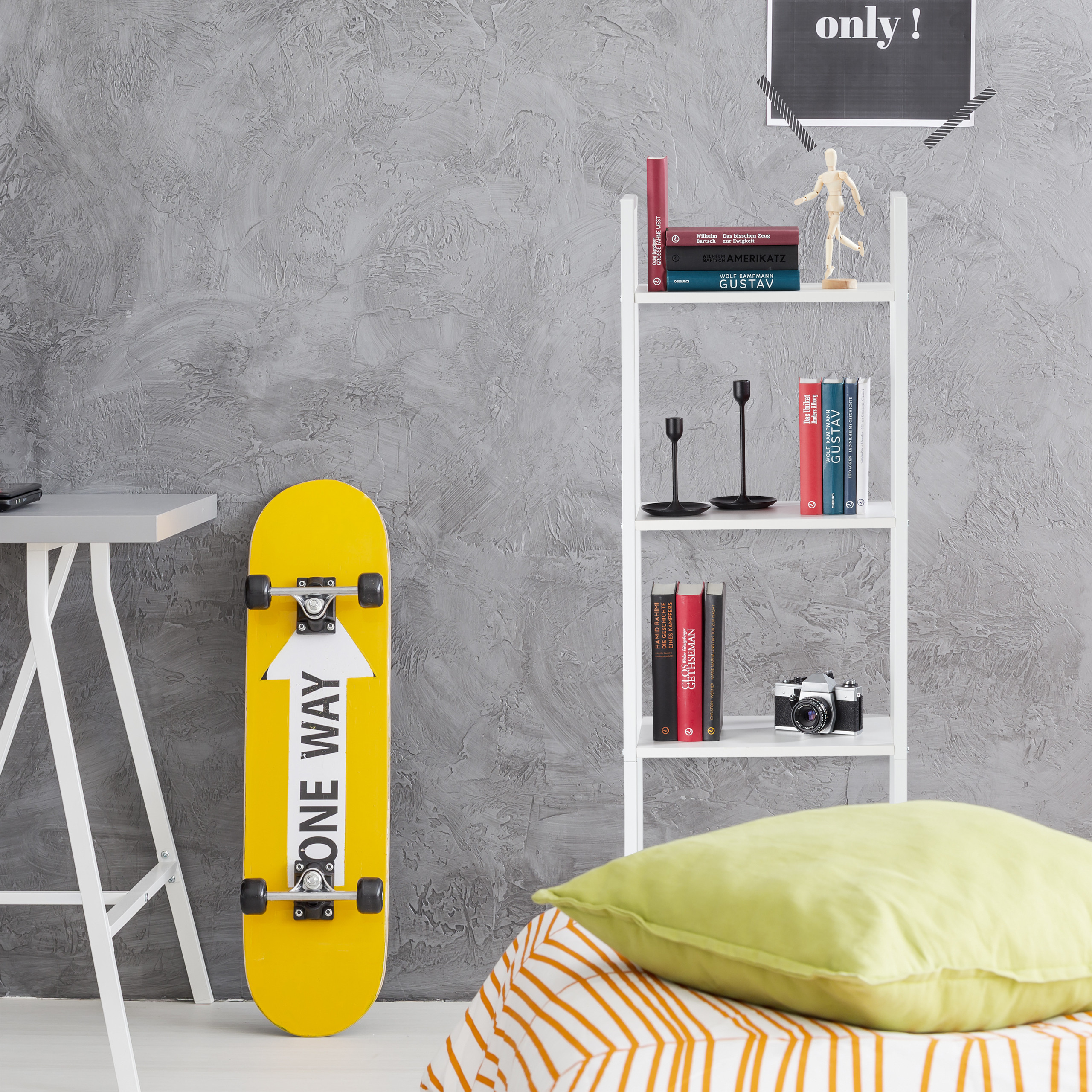 Ein gelbes Skateboard mit „ONE WAY“ lehnt an einer grauen Wand neben einem weißen Bücherregal.