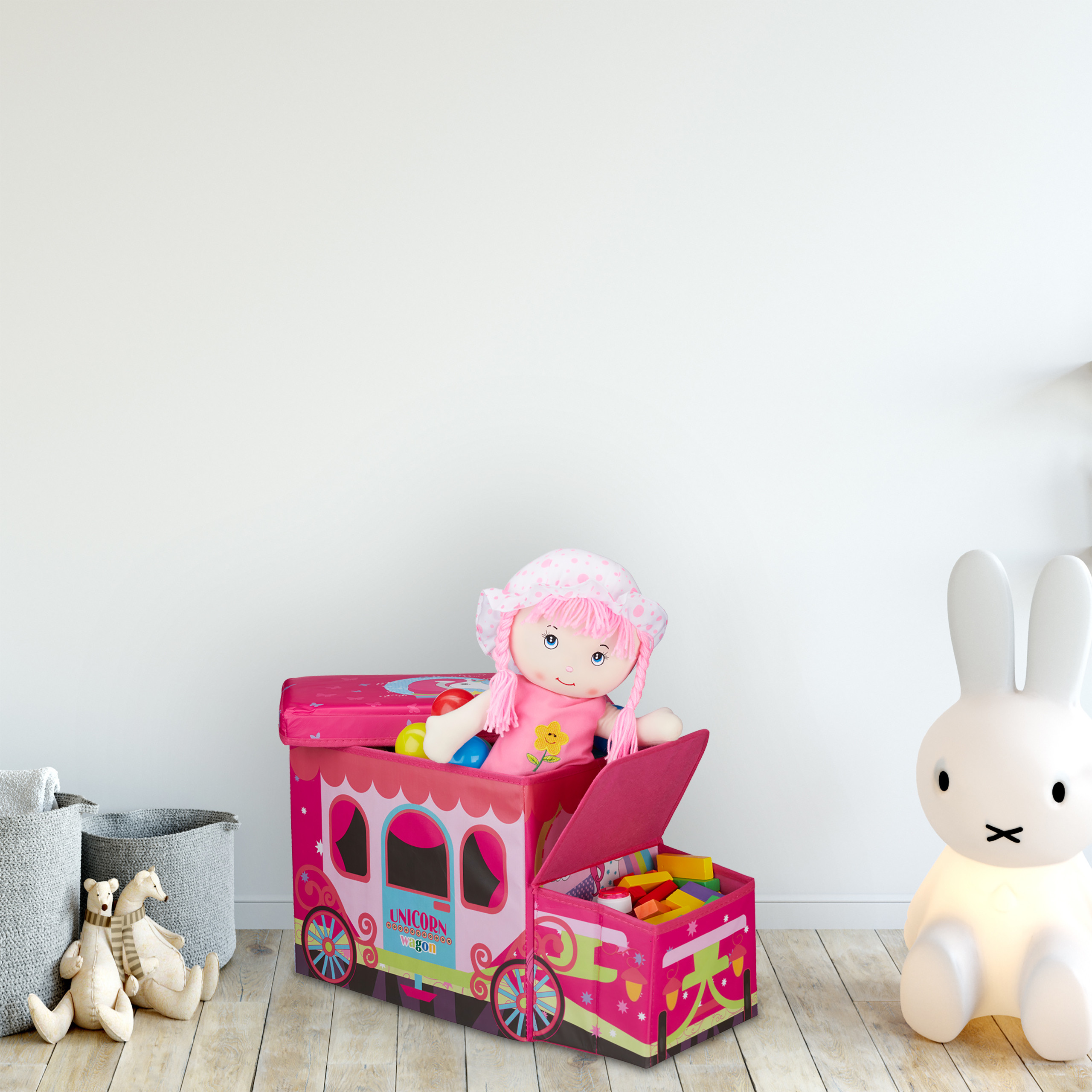 Rosa Spielzeugkiste mit Puppe, Holzklötzen, Stoffbären und einer weißen Hasen-Lampe vor weißer Wand.
