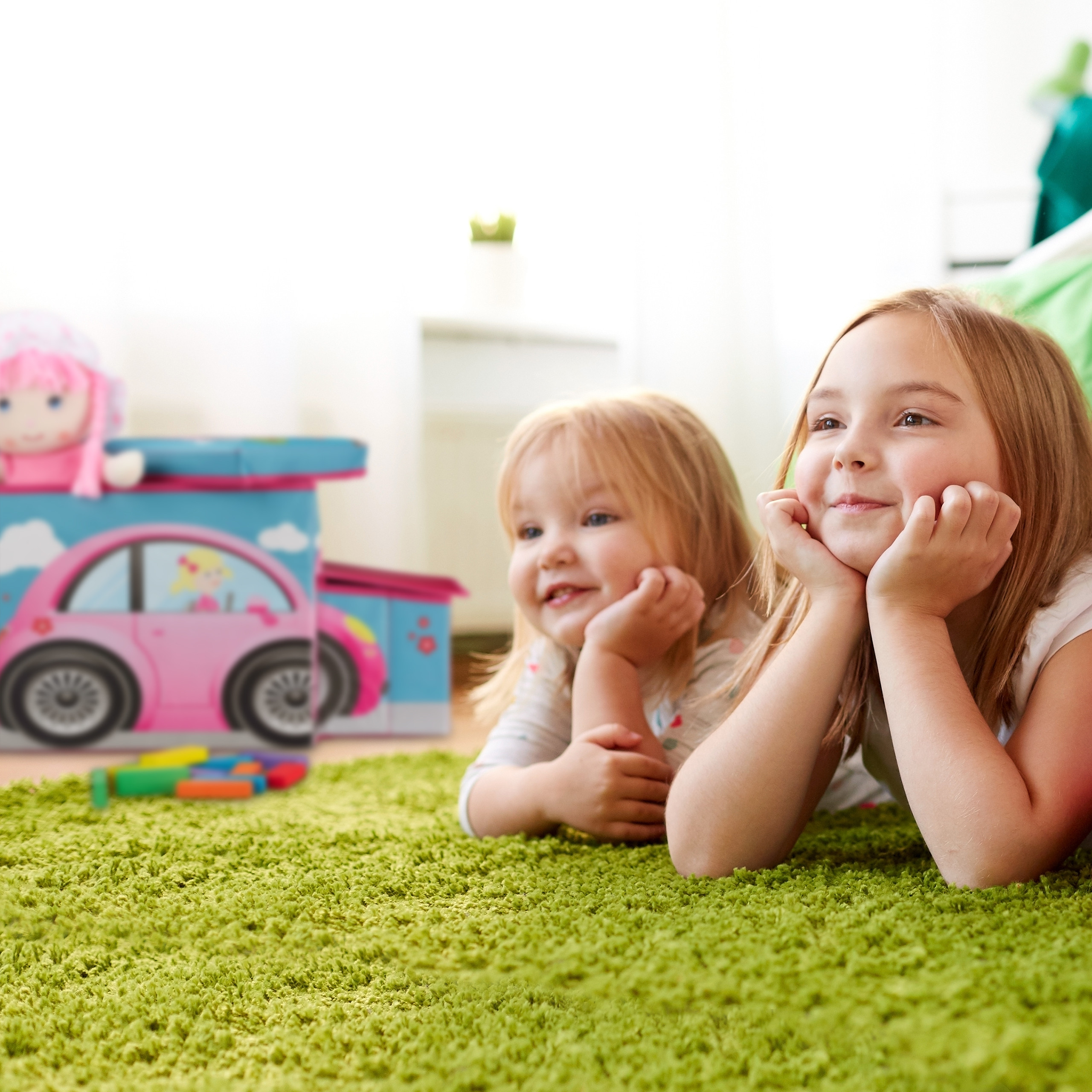 Zwei junge Mädchen liegen auf einem grünen Teppich und stützen das Kinn auf die Hände. Im Hintergrund ein pinkes Spielzeugauto.
