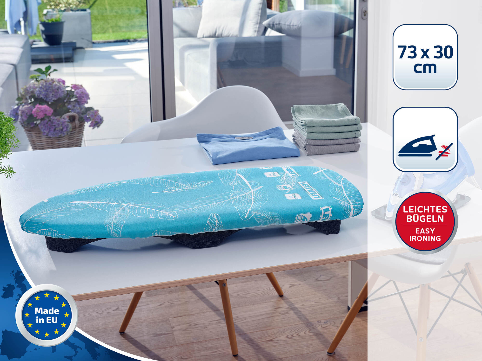 Ein blauer Tisch-Bügeltisch mit weißem Blattmuster liegt auf einem weißen Tisch, daneben gefaltete Wäsche.