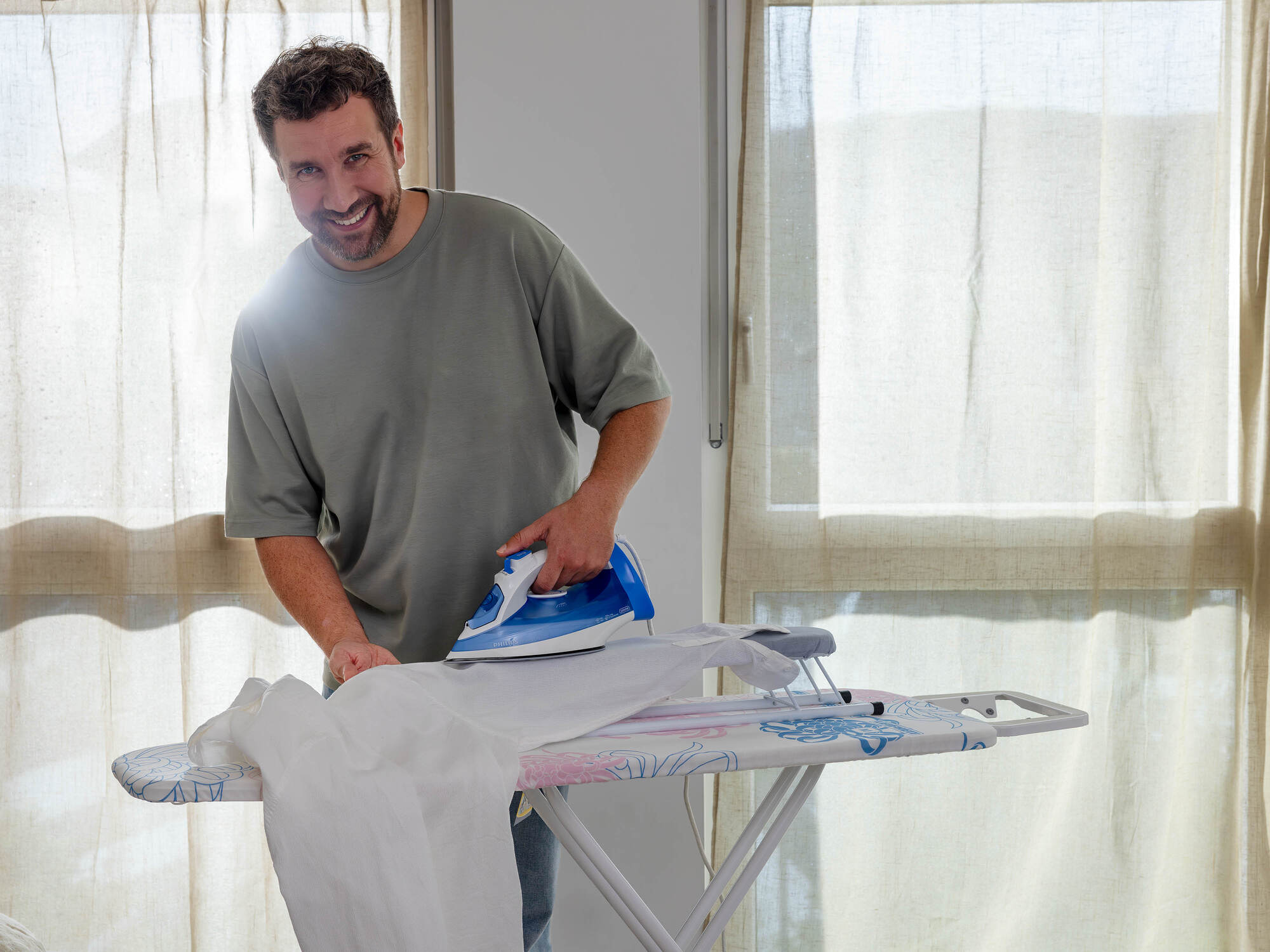 Un uomo sorridente stira abiti bianchi su un'asse da stiro davanti a una finestra luminosa.