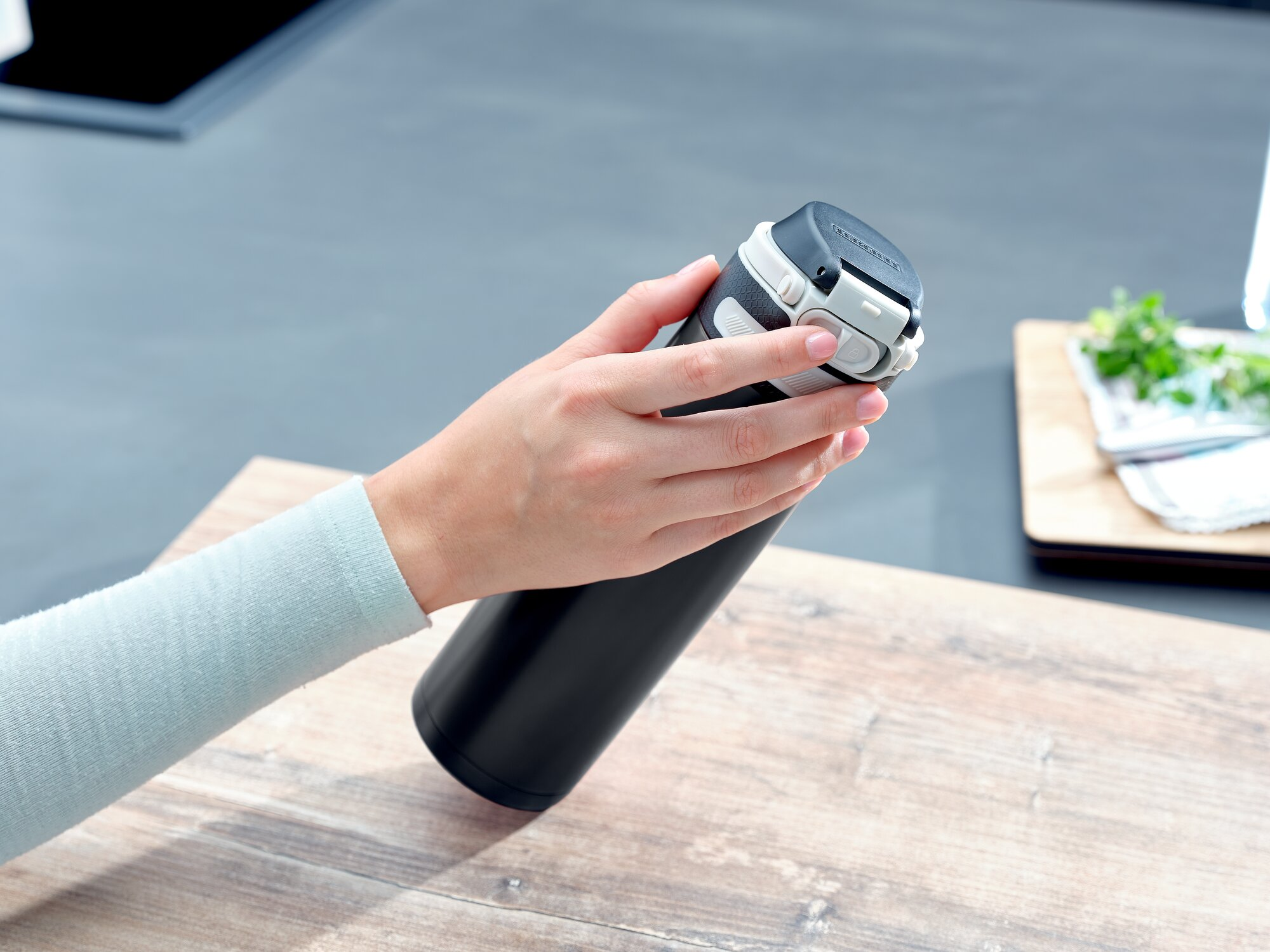 Eine Hand hält eine schwarze Thermosflasche auf einem Holztisch.