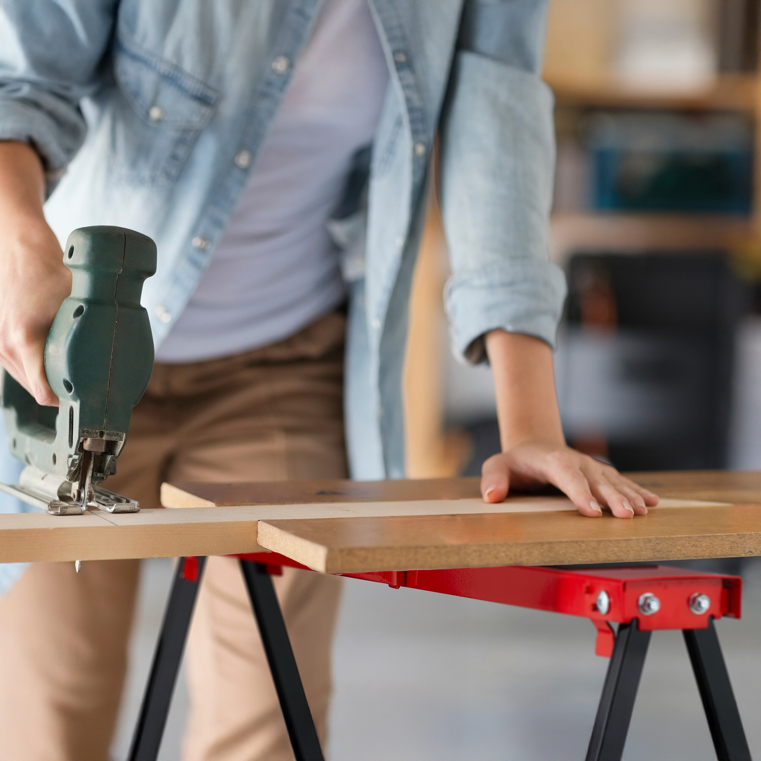 Eine Person schneidet mit einer grünen Stichsäge ein Holzbrett auf einer rot-schwarzen Werkbank.