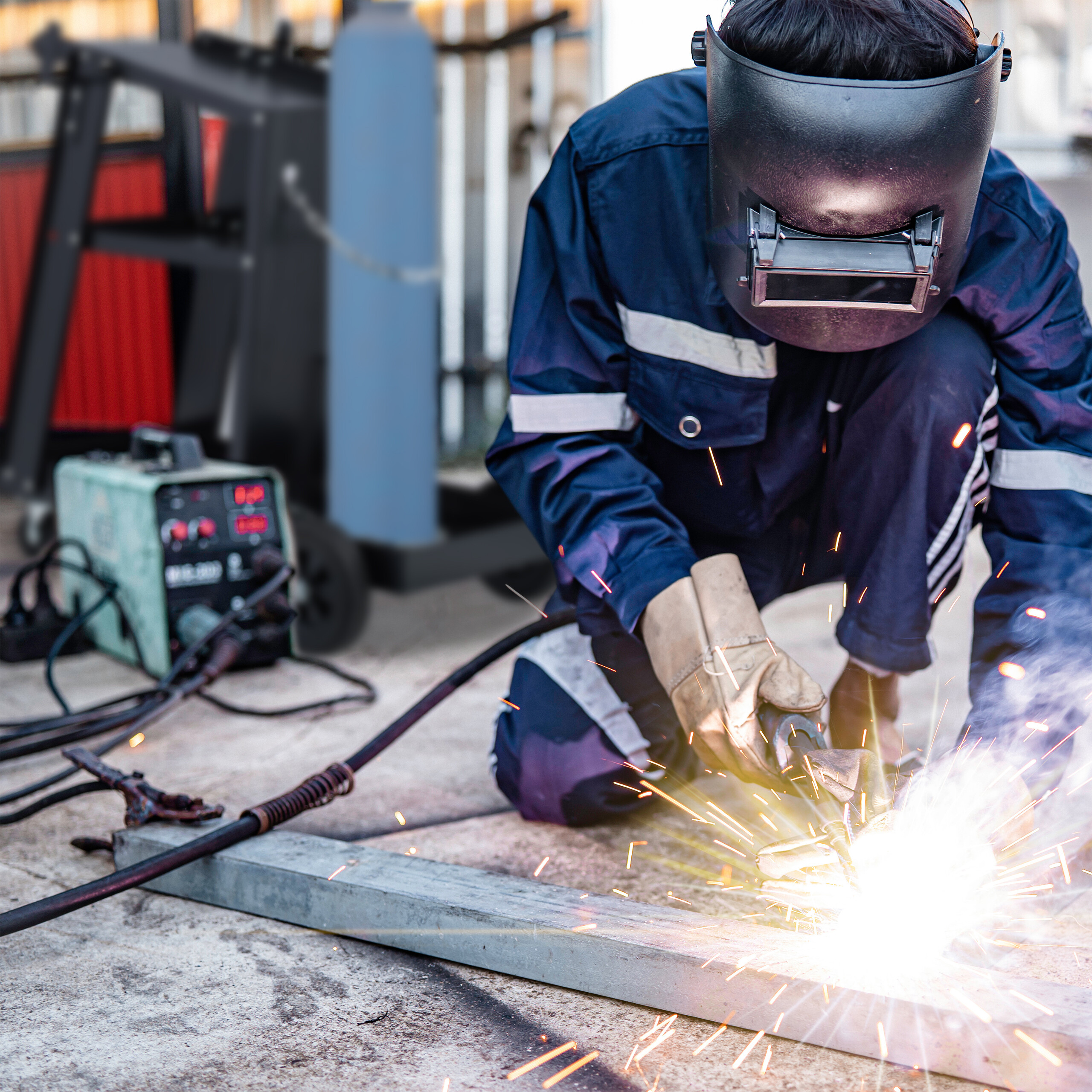 Ein Arbeiter in blauer Schutzkleidung und Schutzhelm schweißt ein Metallstück im Freien, helle Funken fliegen.