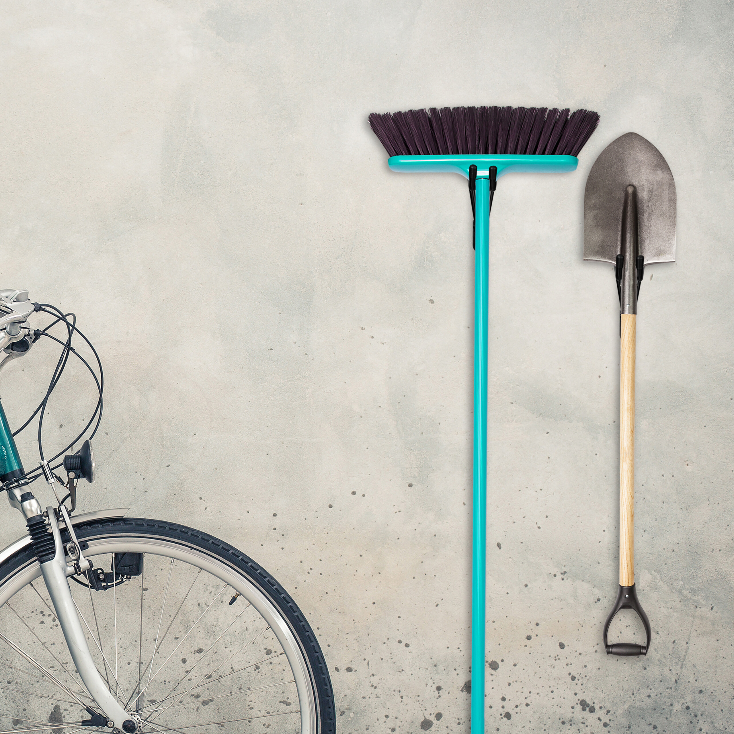 Ein blaues Fahrrad, ein Besen mit türkiser Bürste und eine Schaufel hängen an einer grauen Wand.