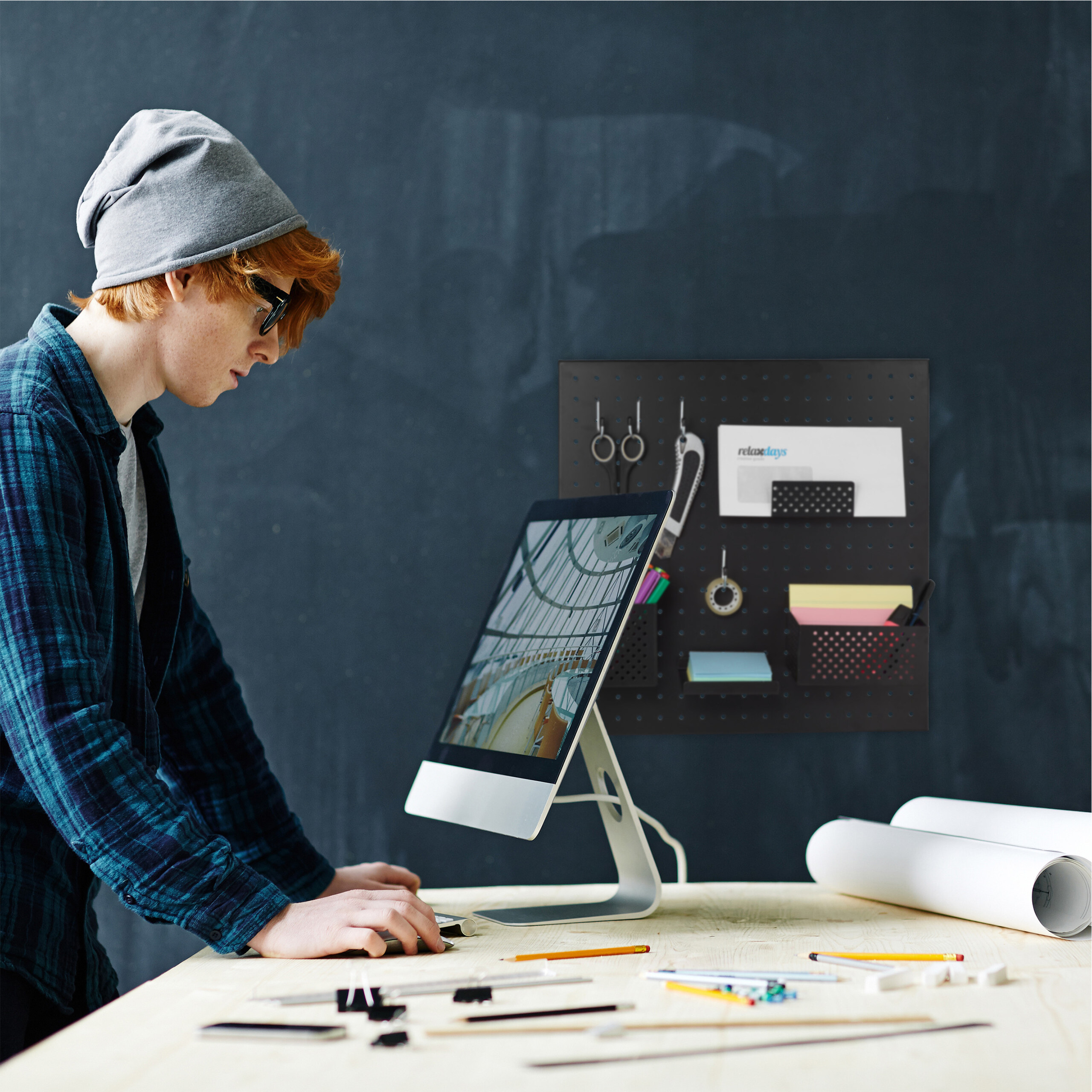 Ein Designer mit Mütze arbeitet an einem Computer an einem Holztisch mit Werkzeugen und Plänen.