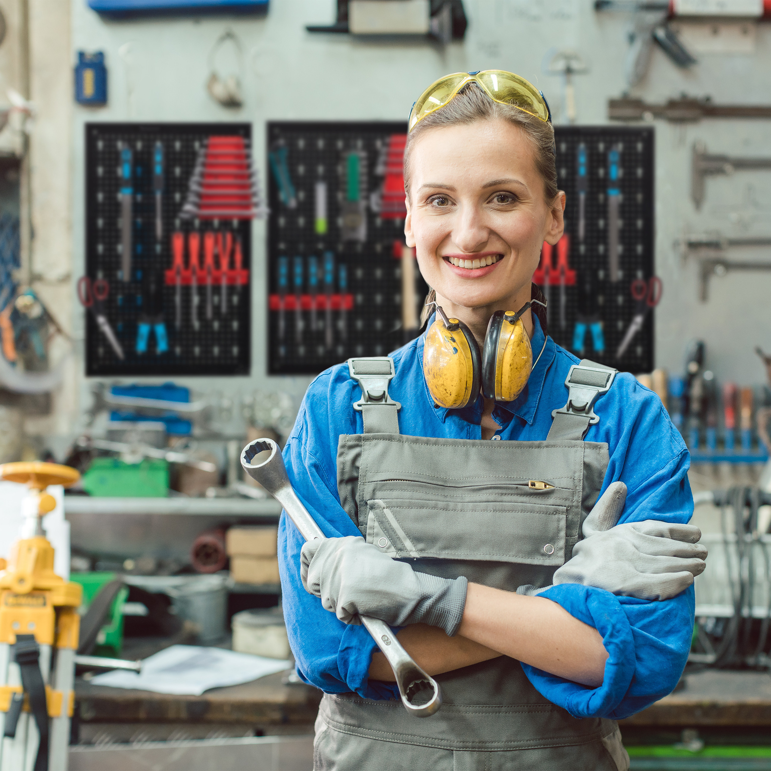 Handwerkerin in blauem Hemd und grauer Latzhose lächelt mit einem Schraubenschlüssel und Gehörschutz in einer Werkstatt.