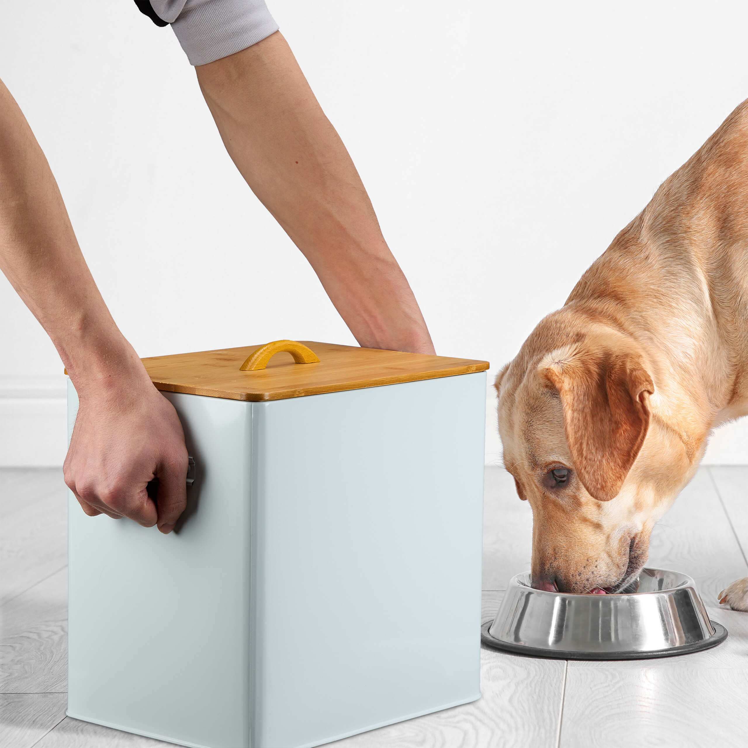Eine Person hält einen hellblauen Behälter mit Holzdeckel, während ein Golden Retriever aus einer Metallschüssel frisst.