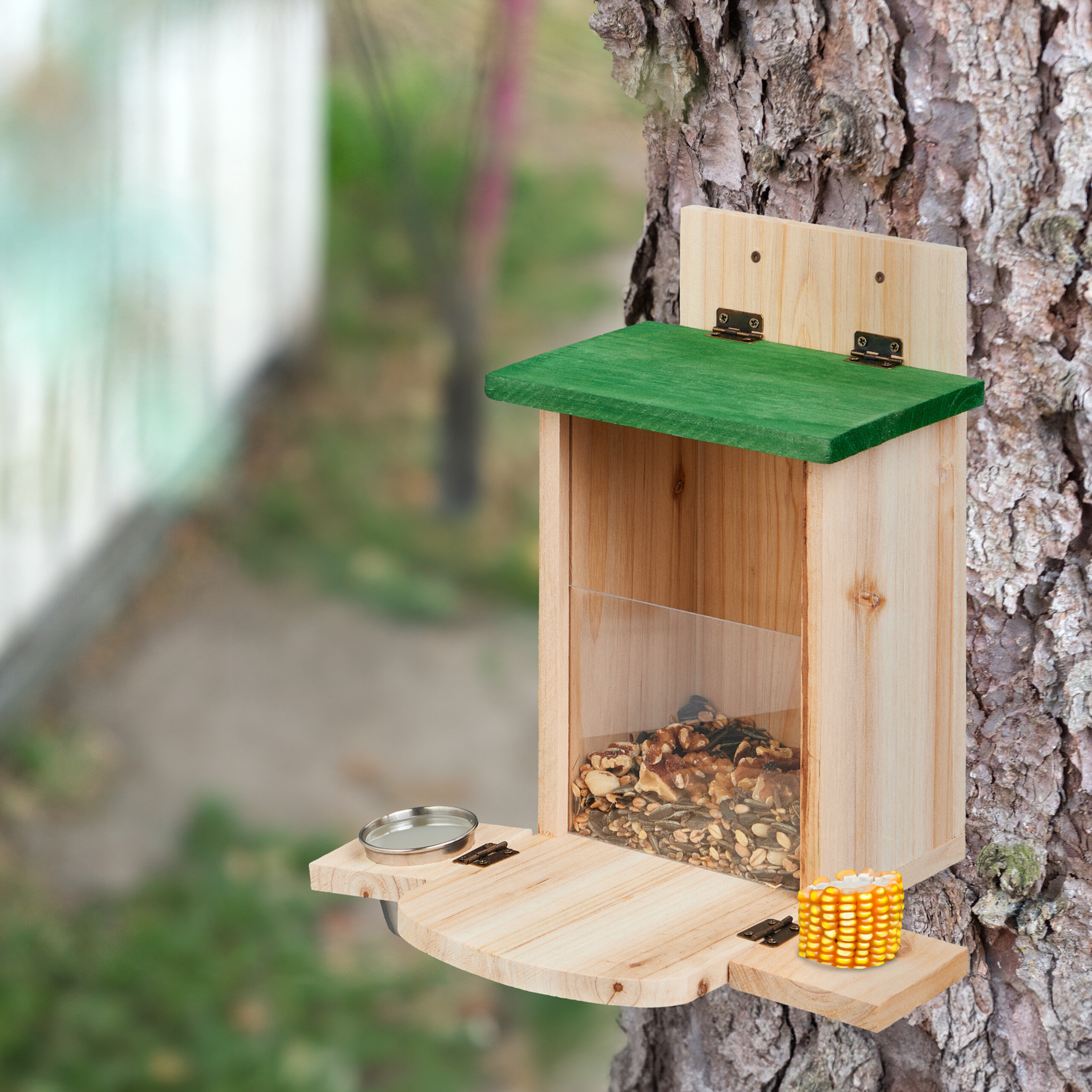 Holzvogelhäuschen mit grünem Dach, Vogelfutter und Maiskolben an einem Baum.