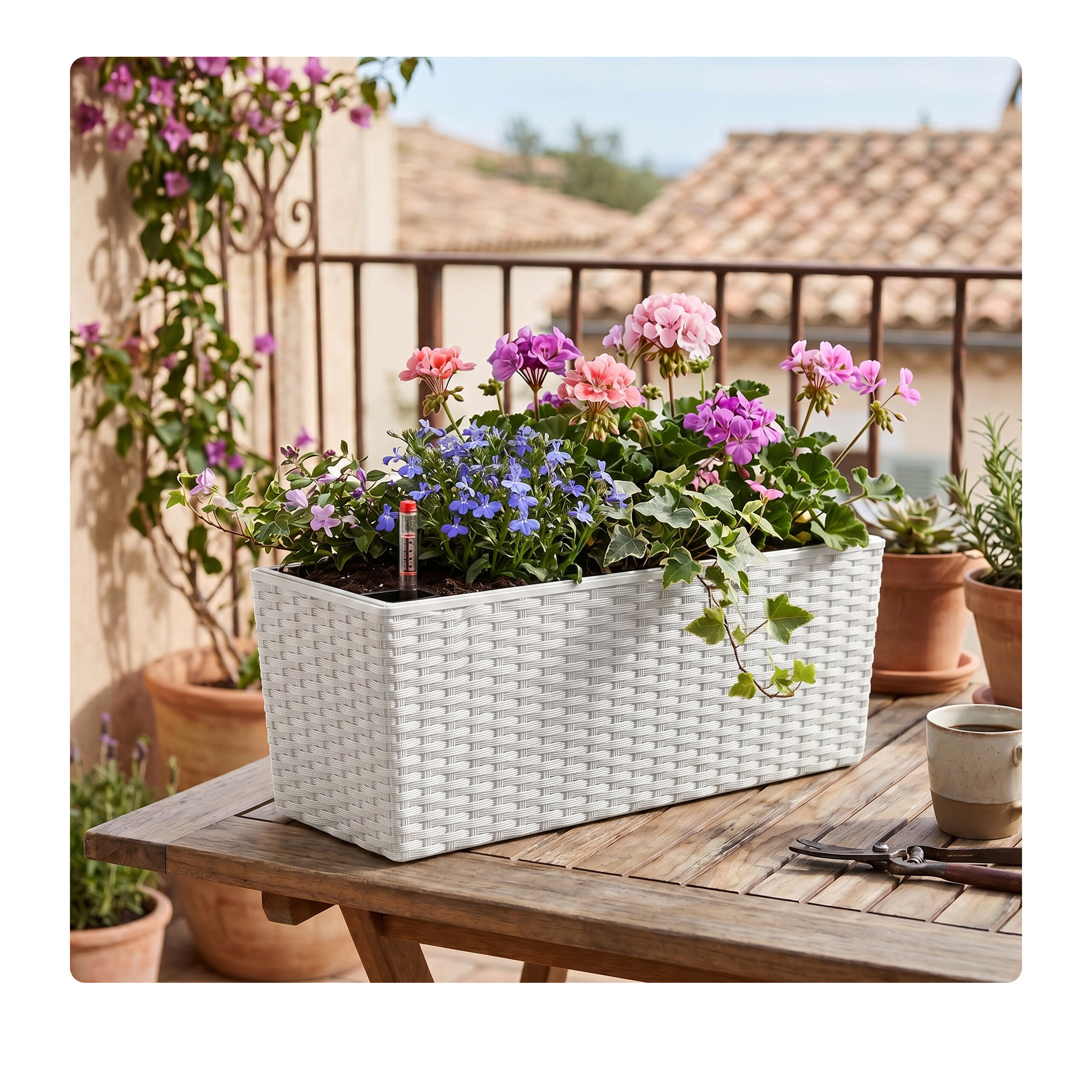 Ein weißer Rattankasten mit bunten Blumen und Efeu steht auf einem Holztisch auf einem Balkon.