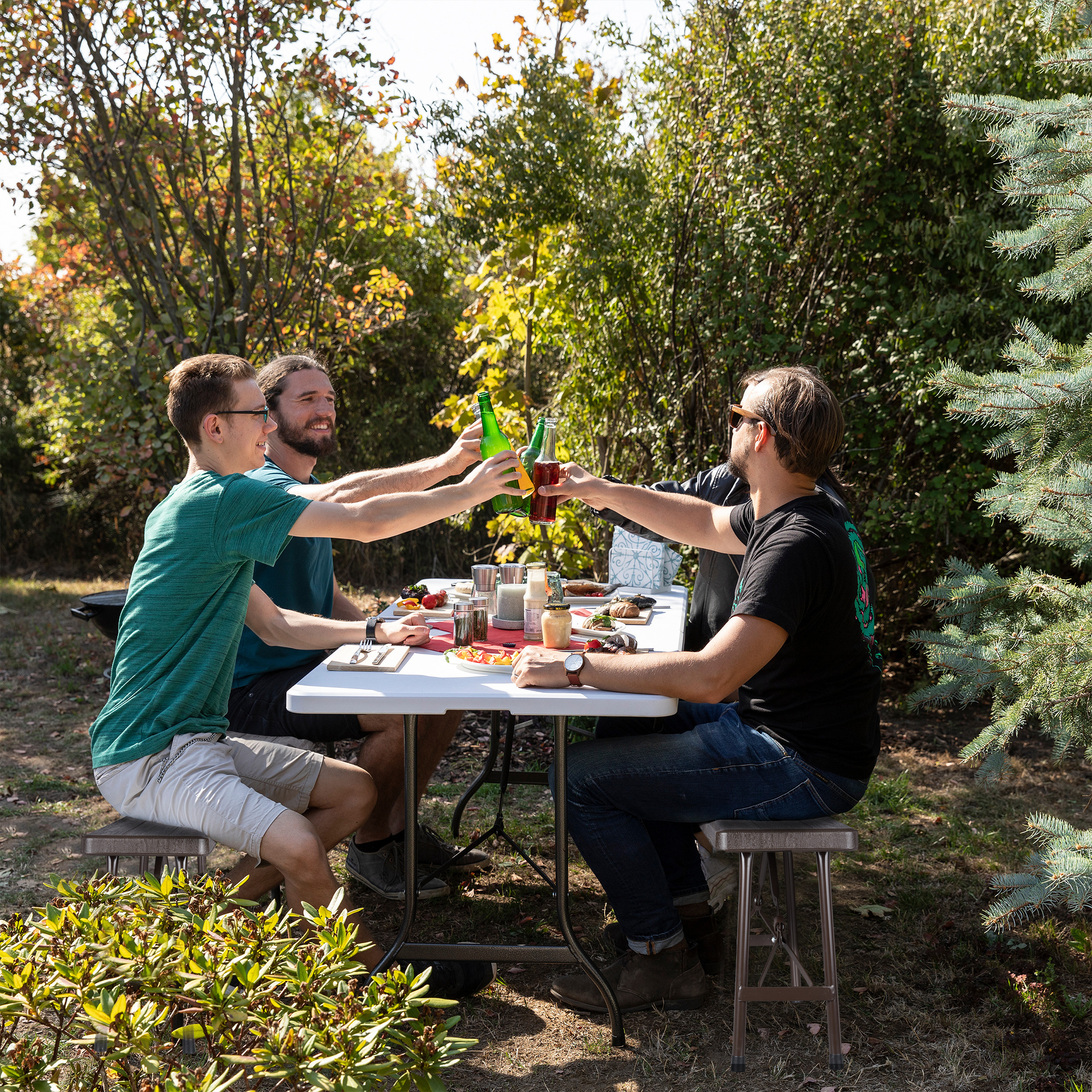 Drei Männer sitzen an einem Gartentisch und stoßen in einem sonnigen Garten mit Bierflaschen an.