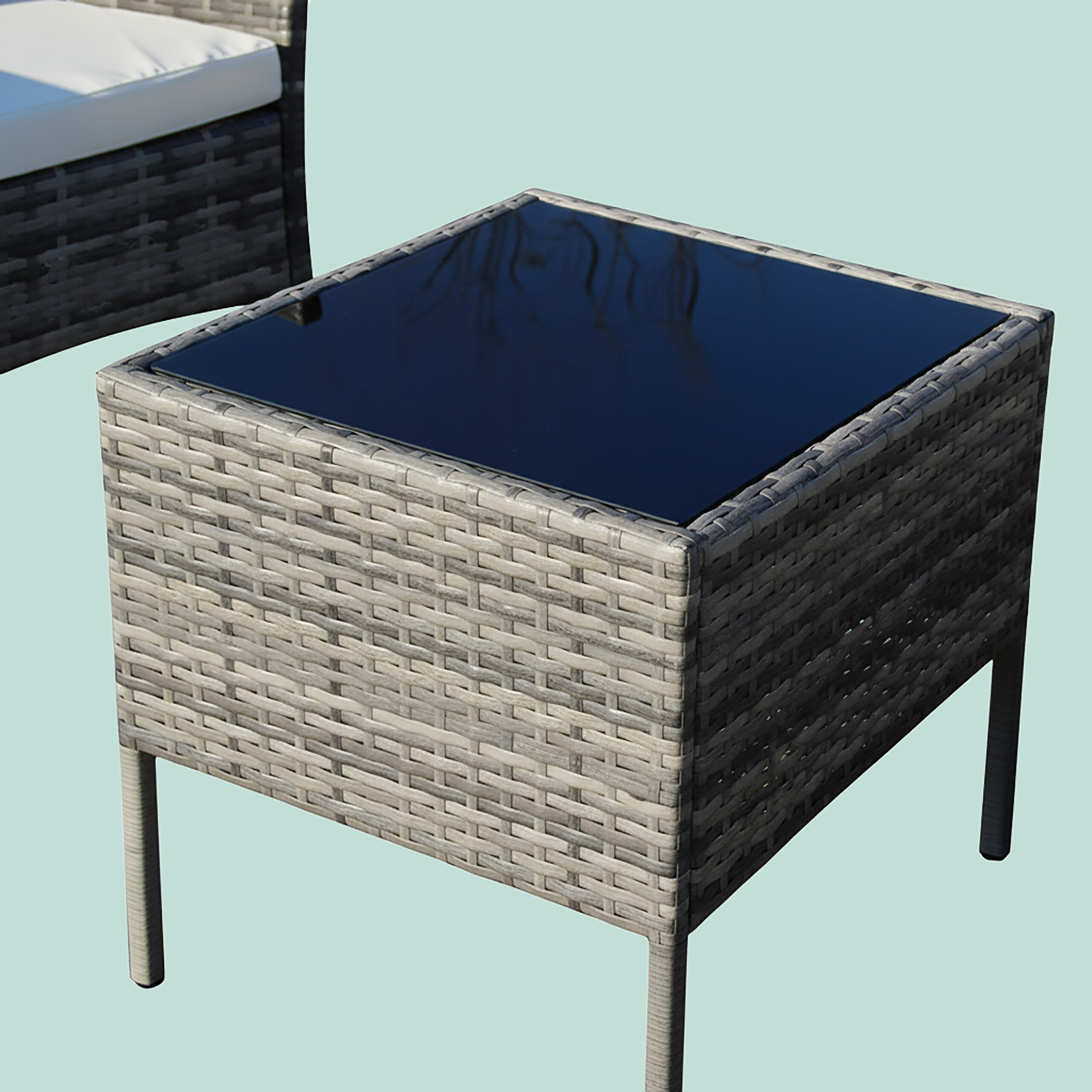 Ein quadratischer Rattan-Beistelltisch mit schwarzer Glasplatte steht vor einem hellblauen Hintergrund.