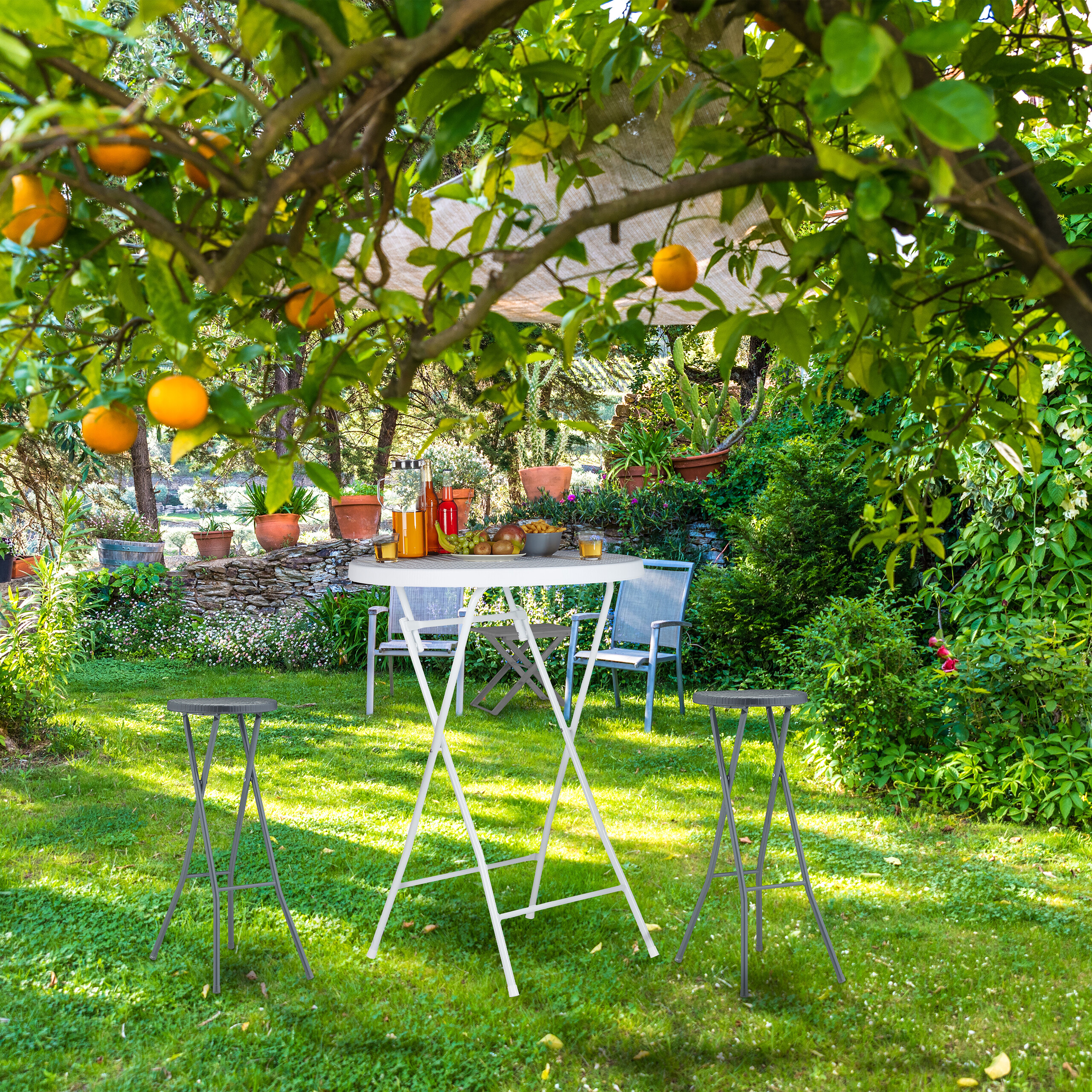 Ein weißer Tisch mit Getränken und Hocker steht auf einer grünen Wiese unter einem Orangenbaum.
