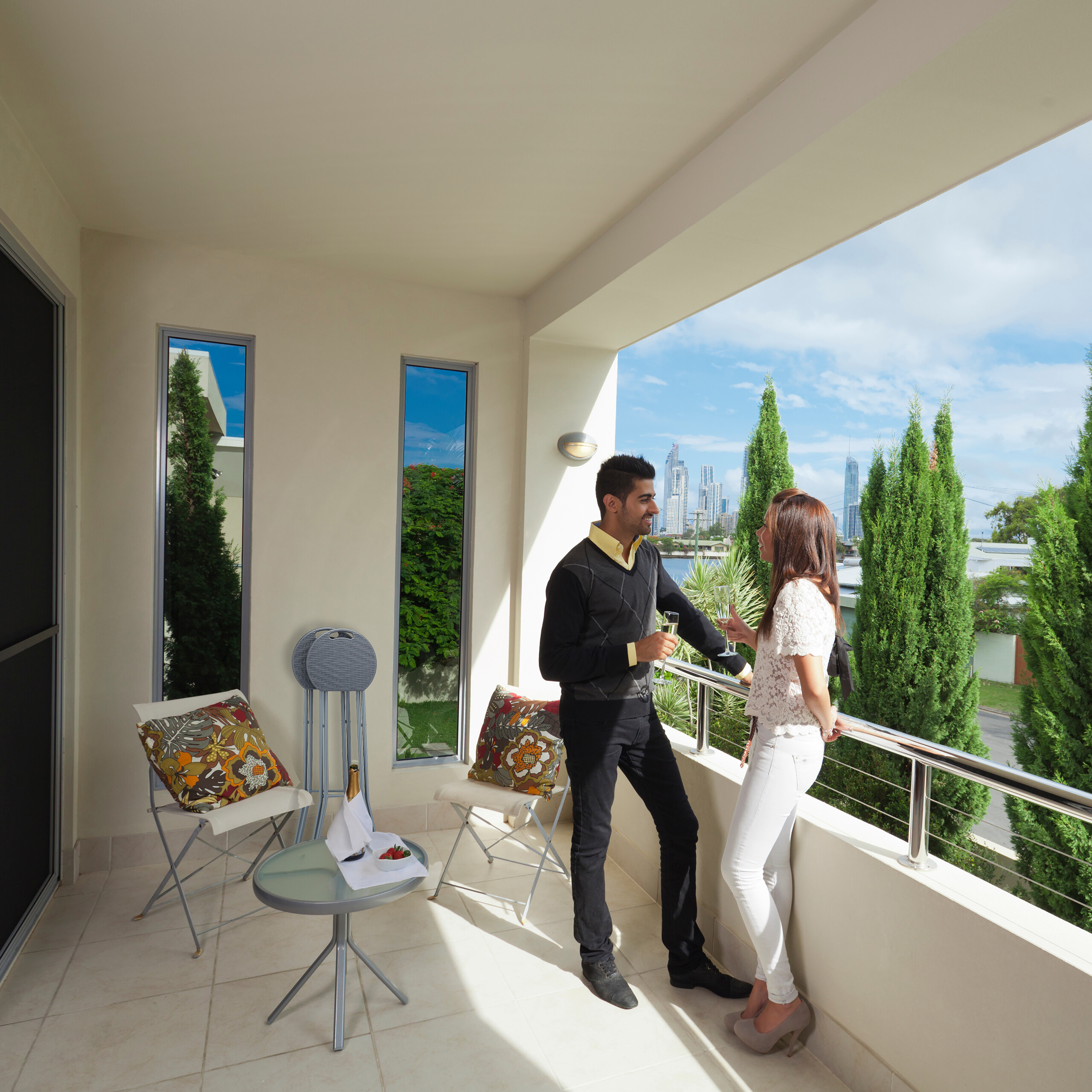 Ein Mann und eine Frau stehen auf einem Balkon mit Champagnergläsern und blicken auf eine Skyline.