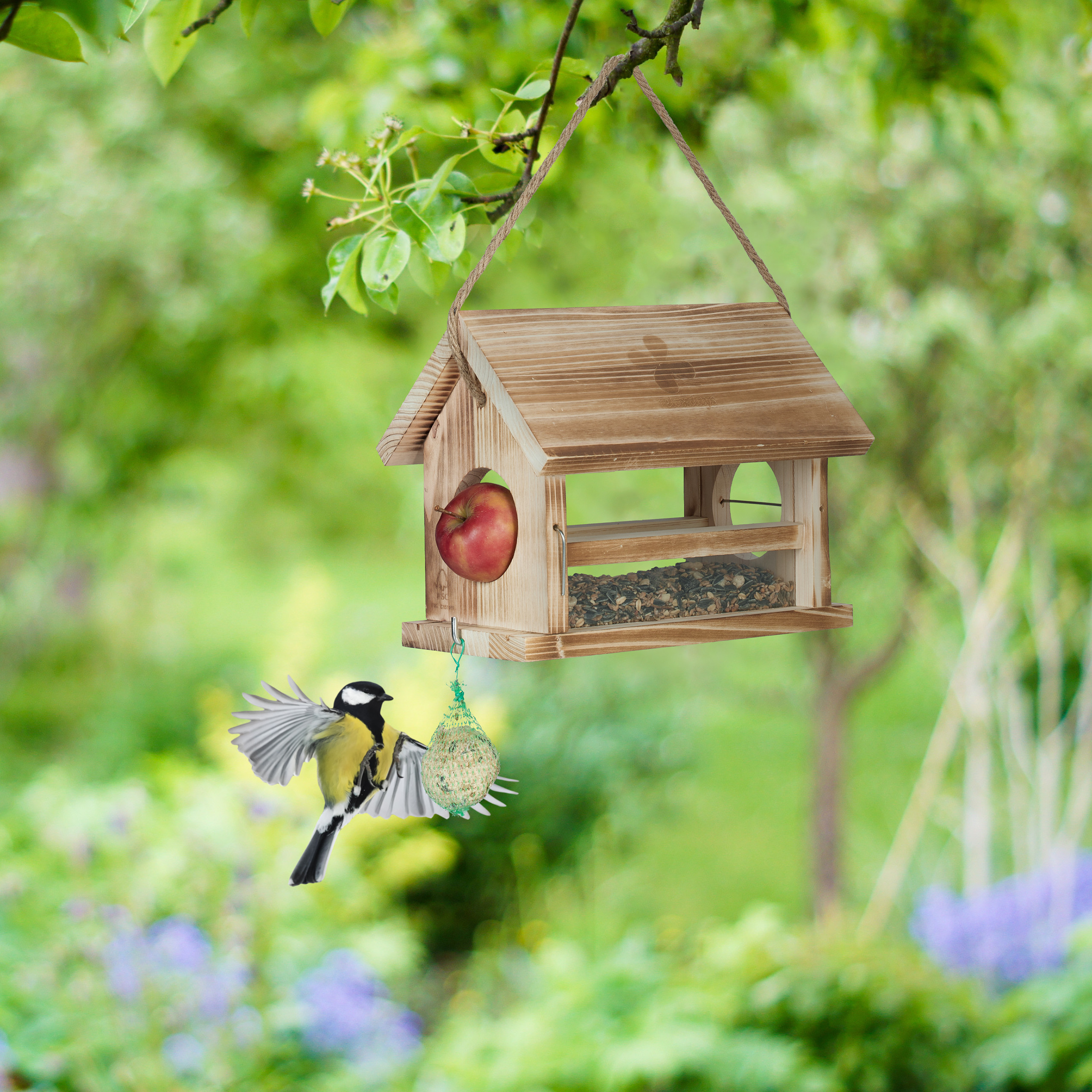 Eine Meise fliegt zu einem hölzernen Vogelnäher mit Apfel und Futter vor grünem Hintergrund.