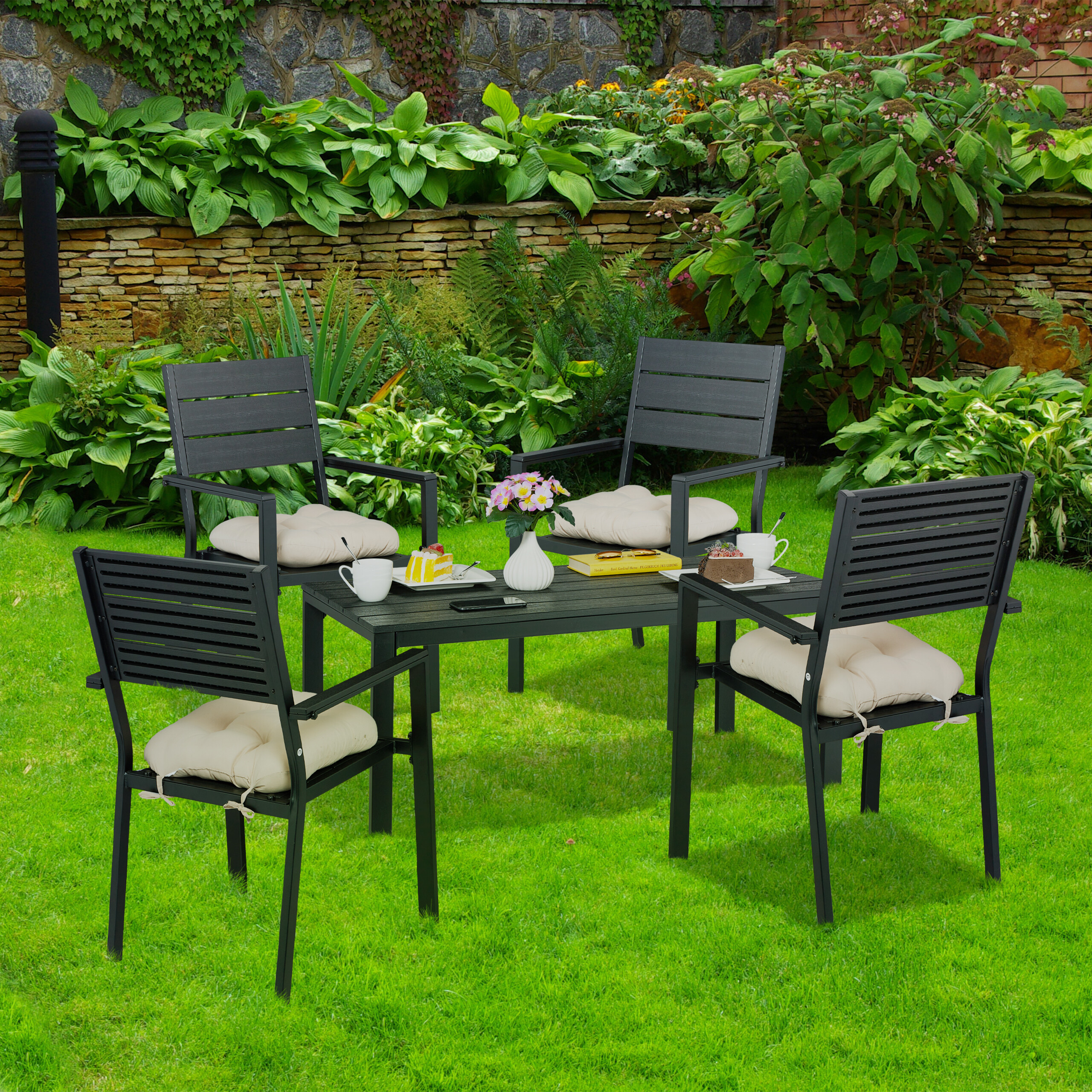 Ein schwarzer Terrassentisch mit vier Stühlen und beigen Kissen auf grünem Rasen vor einer Gartenkulisse.