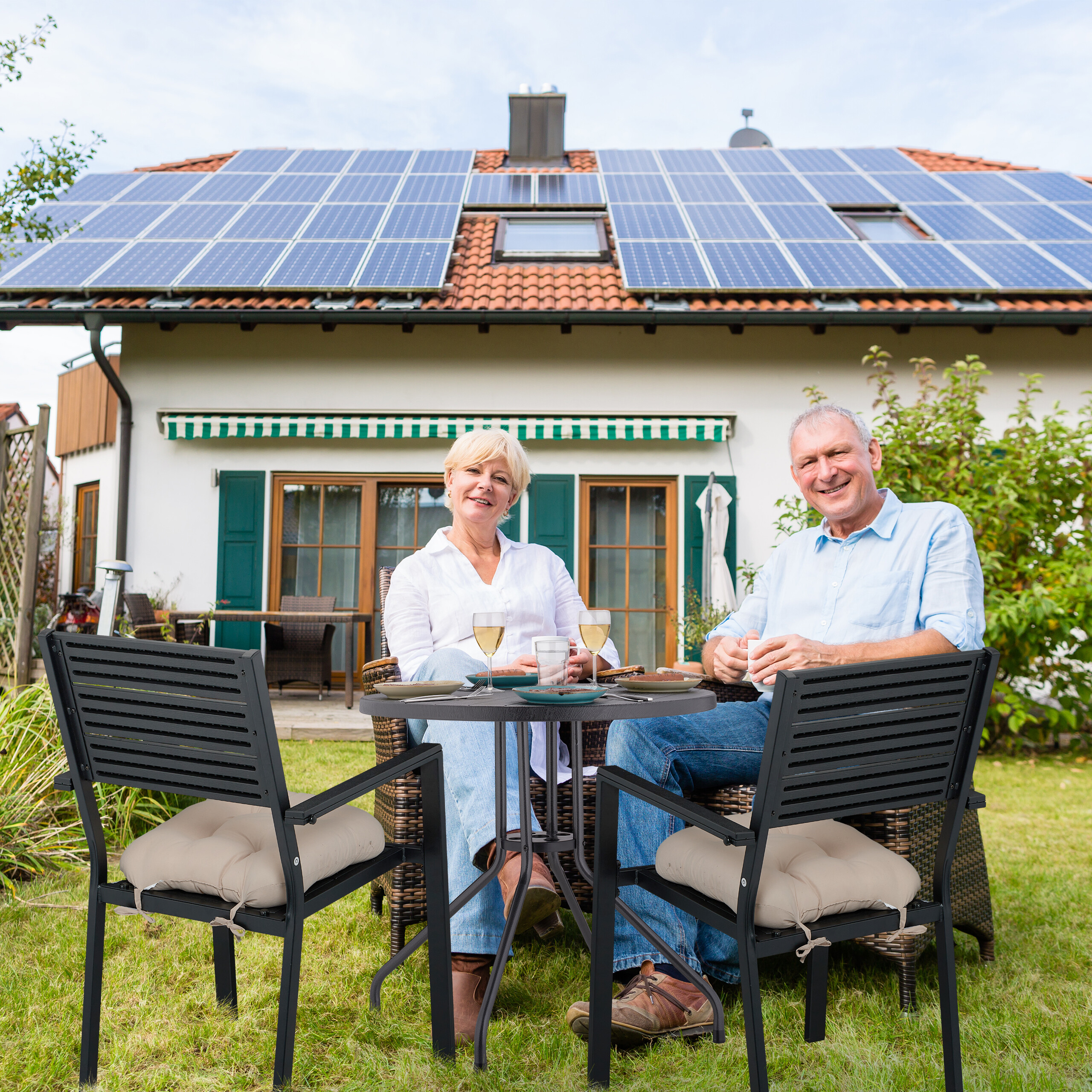Älteres Paar sitzt im Garten vor einem Haus mit Solaranlage.