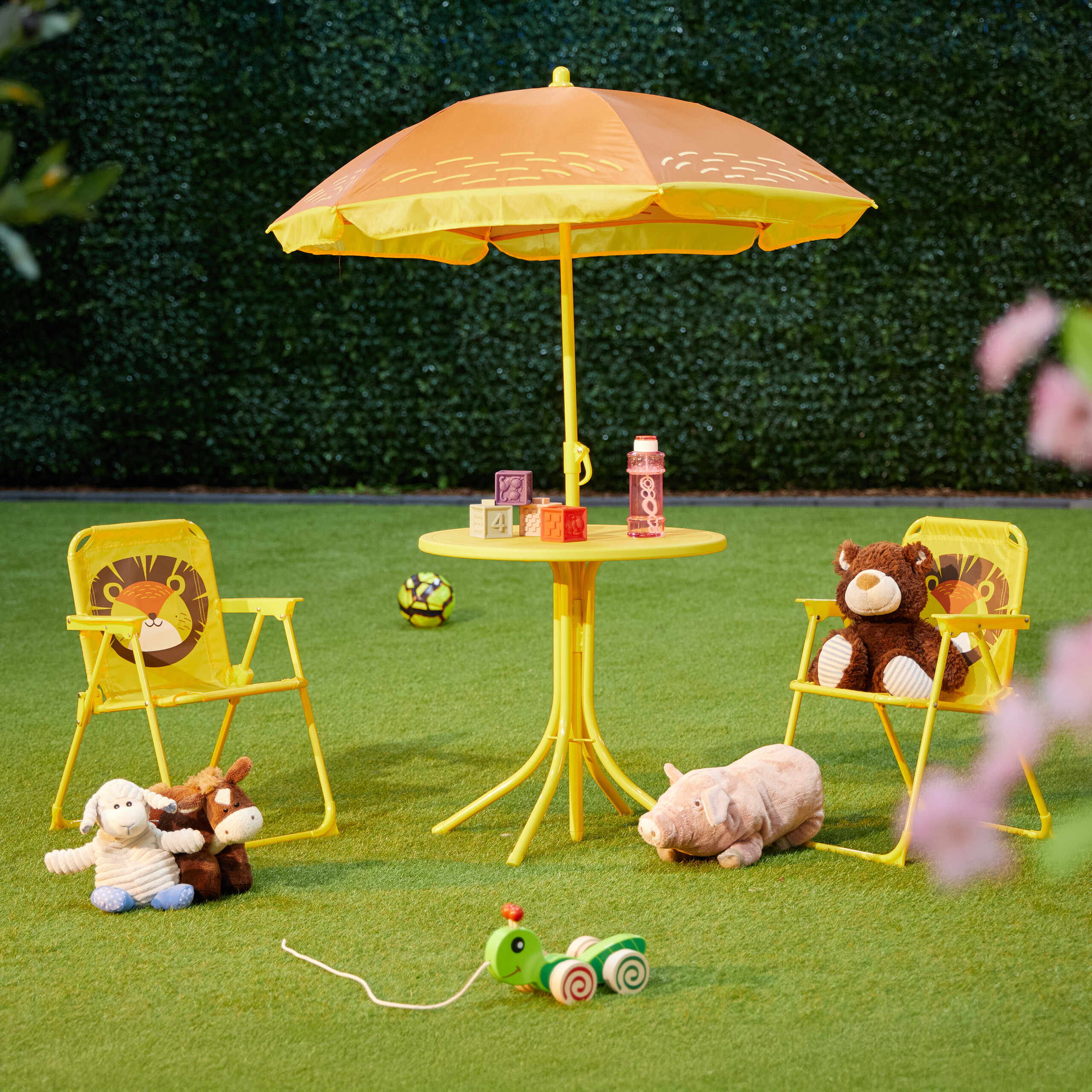 Gelber Kindertisch mit Sonnenschirm, Stühlen, Plüschtieren und Spielzeug auf grünem Rasen vor Hecke.