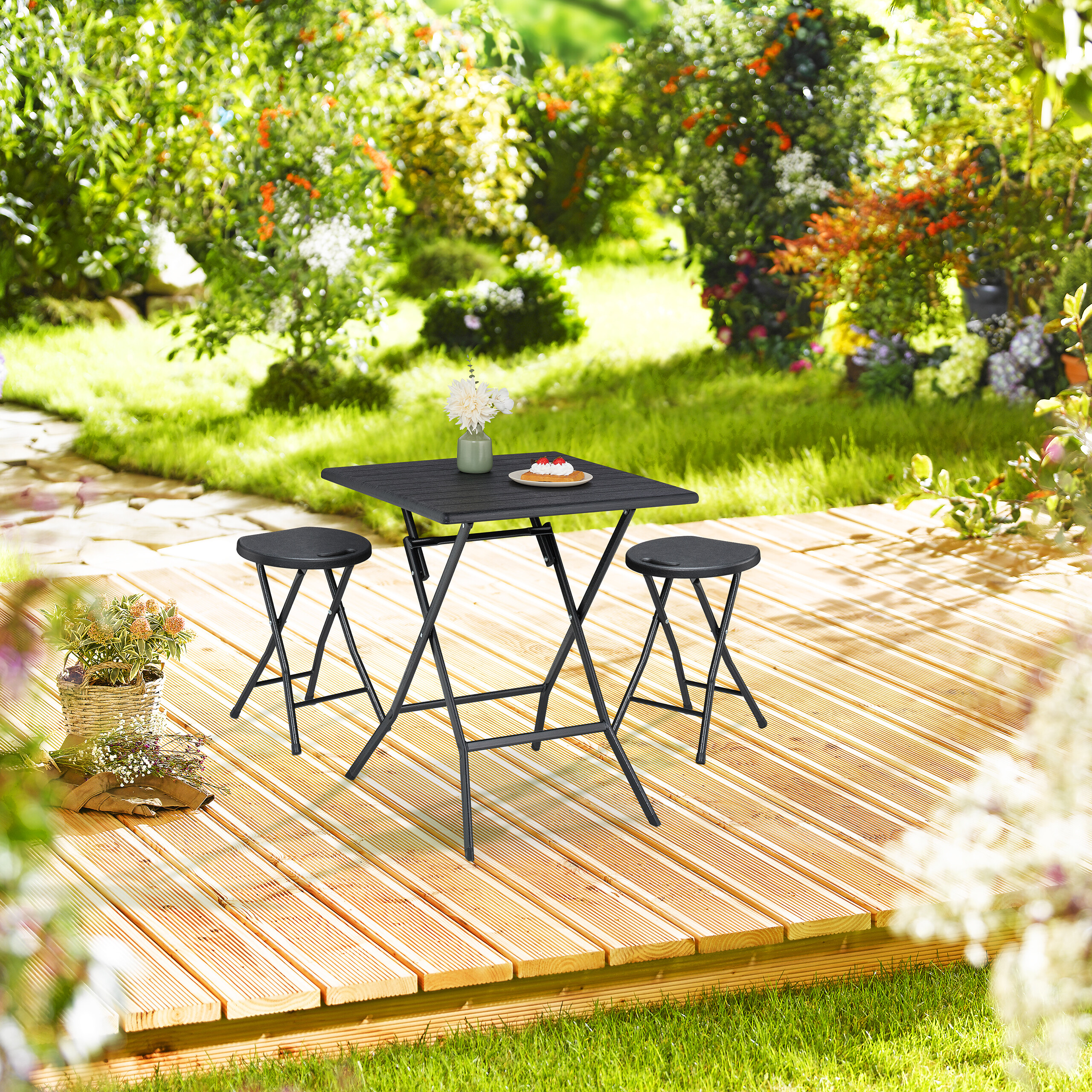 Ein schwarzer Klapptisch mit zwei Hockern steht auf einer Holzterrasse in einem sonnigen, grünen Garten.