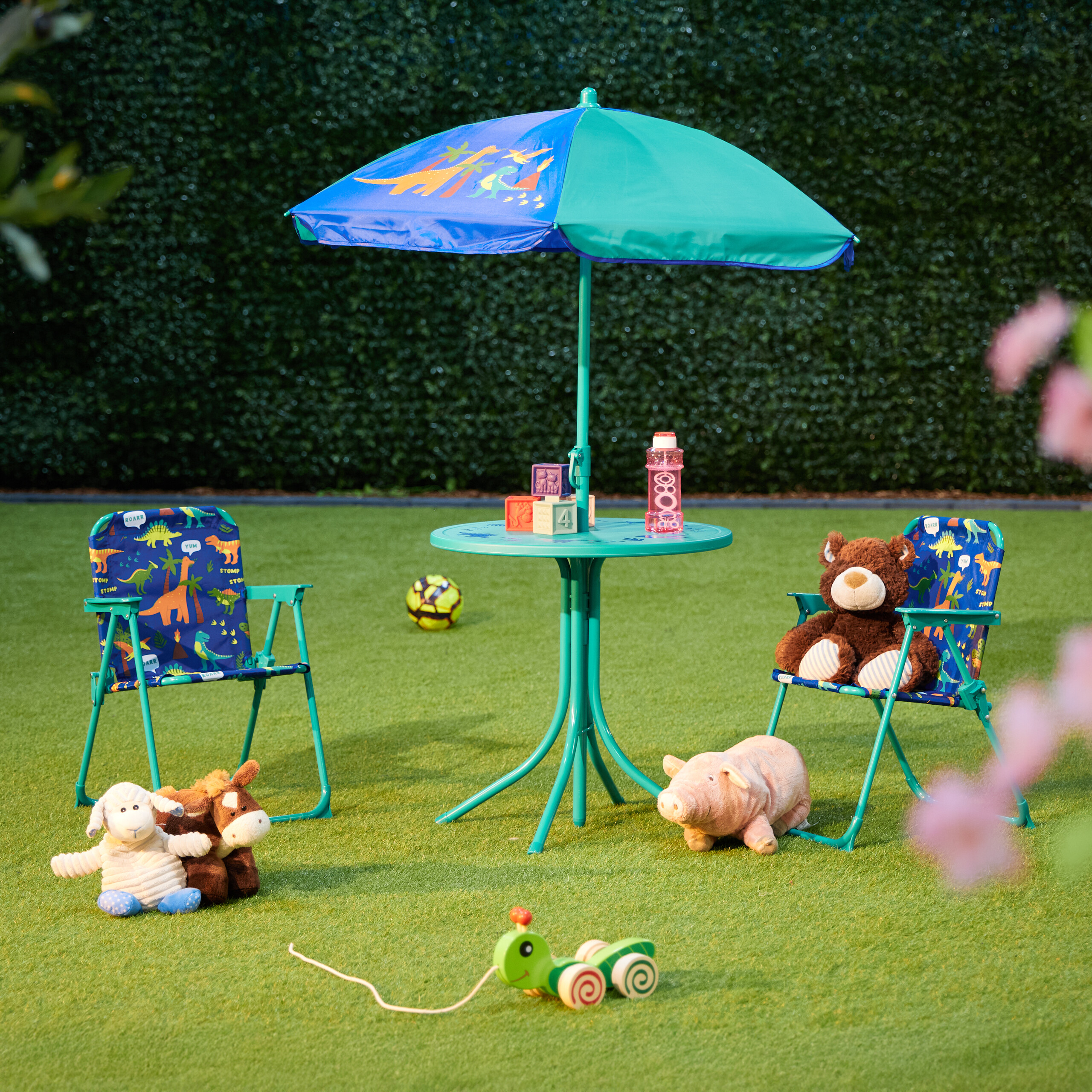 Kindermöbelset mit blau-grünem Regenschirm, zwei Stühlen, Spielzeug, auf grünem Rasen vor Hecke.
