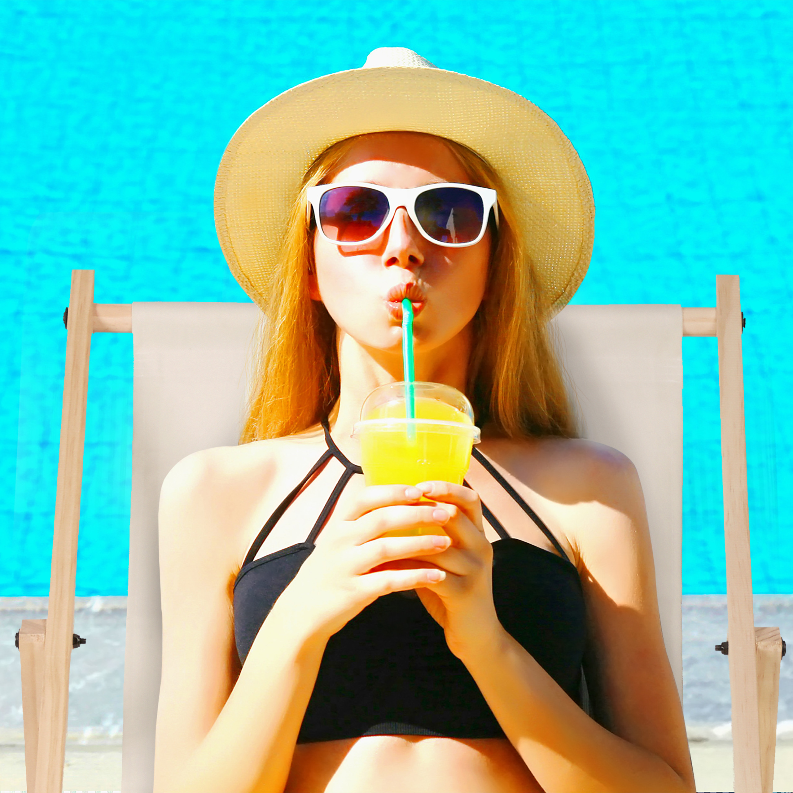 Frau mit Sonnenbrille und Hut sitzt auf einer Liege und trinkt Saft am Pool.