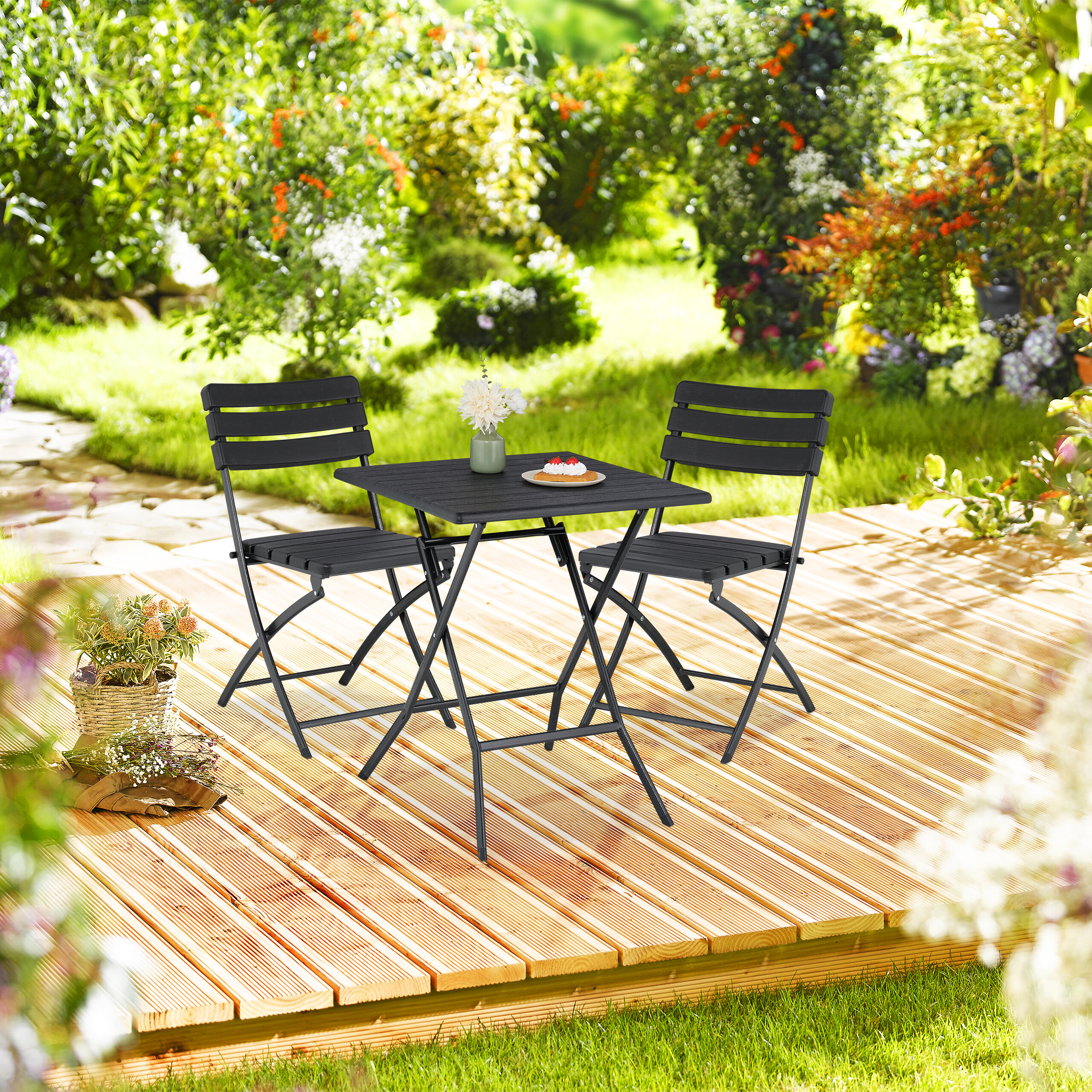 Ein schwarzer Klapptisch mit zwei Stühlen auf einer Holzterrasse im sonnigen Garten.
