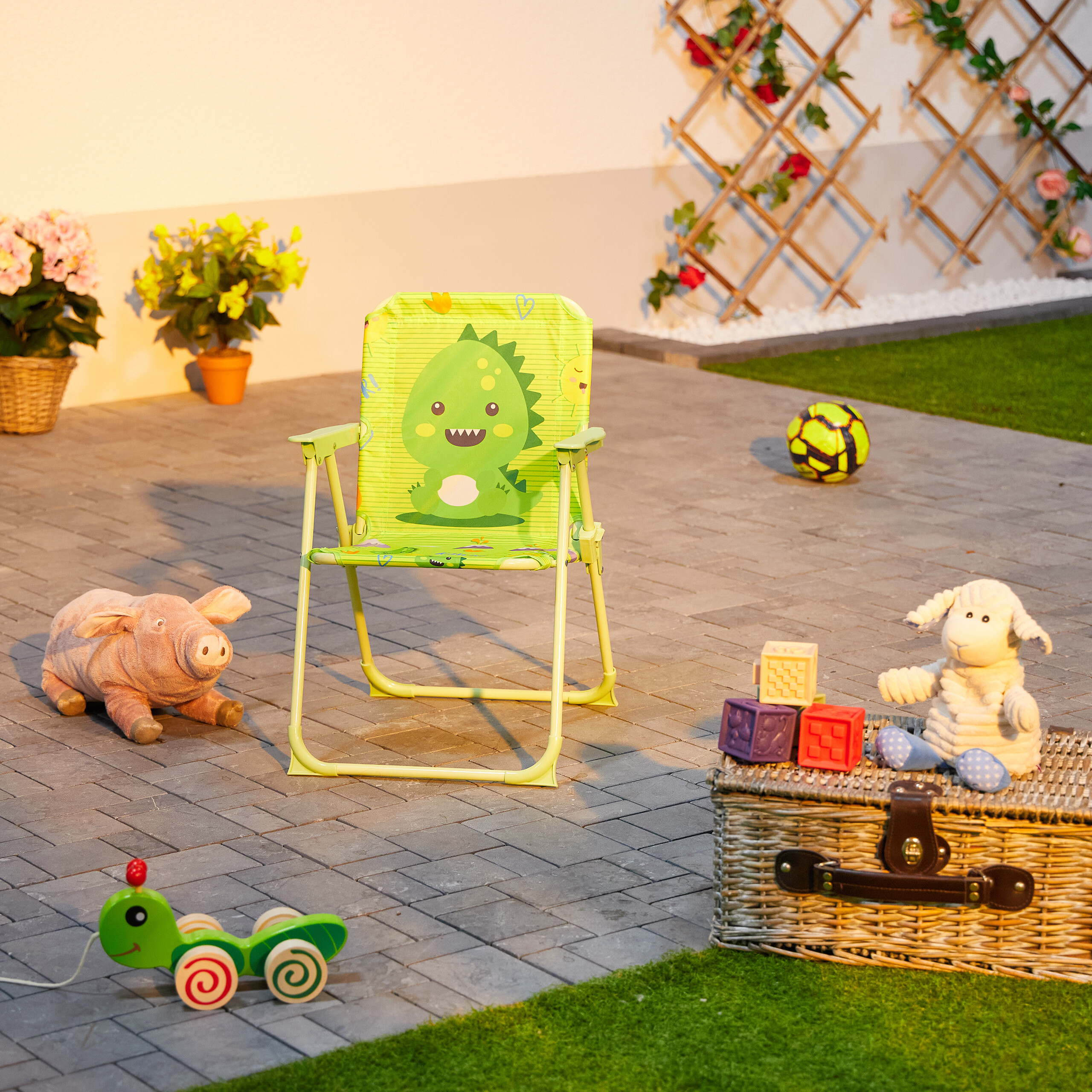 Gruener Kinder-Klappstuhl mit Dino-Motiv auf gepflasterter Terrasse, umgeben von Spielzeug, Blumen und Gras.