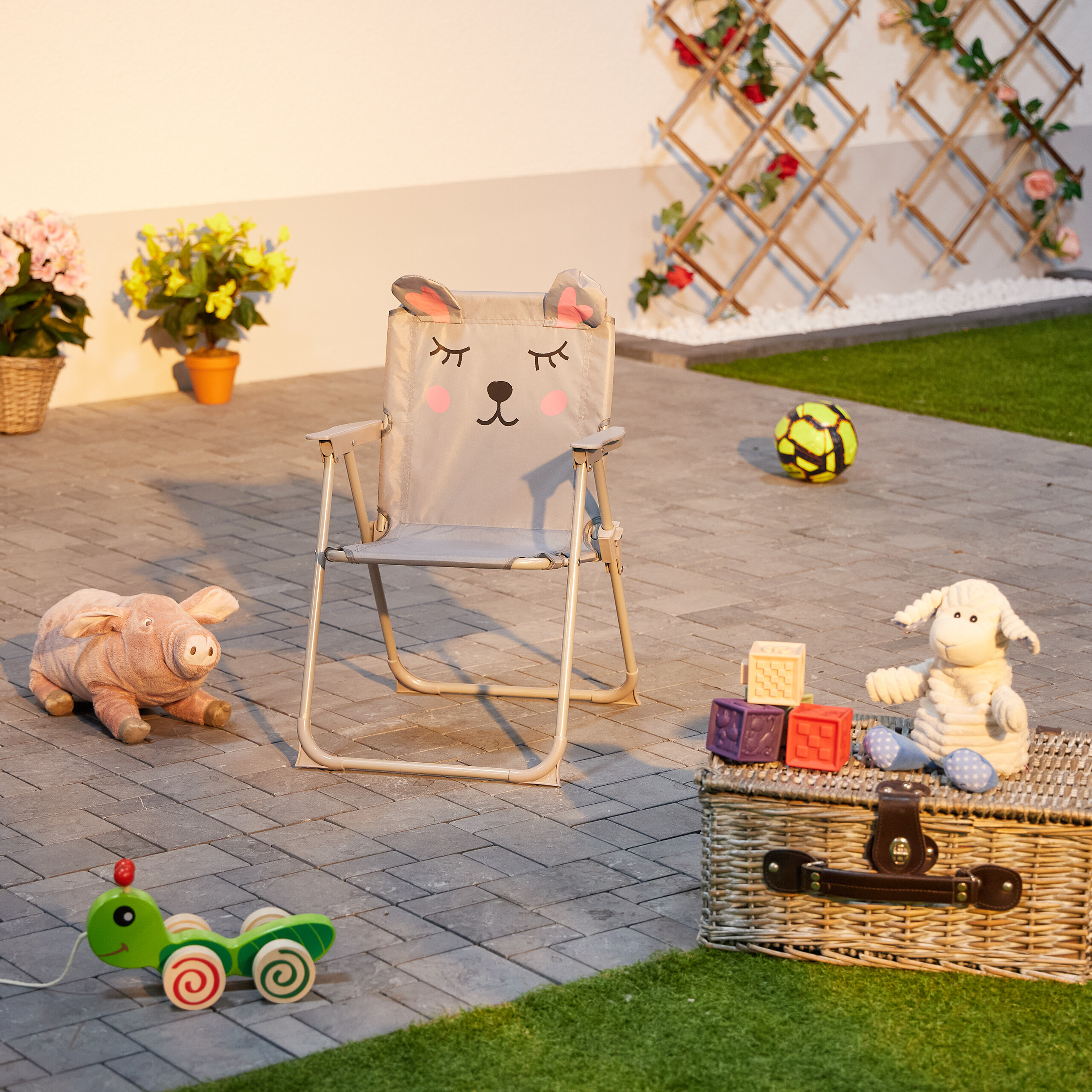 Ein grauer Kinder-Klappstuhl mit Tiermotiv auf Steinen, umgeben von Spielzeug im Garten.