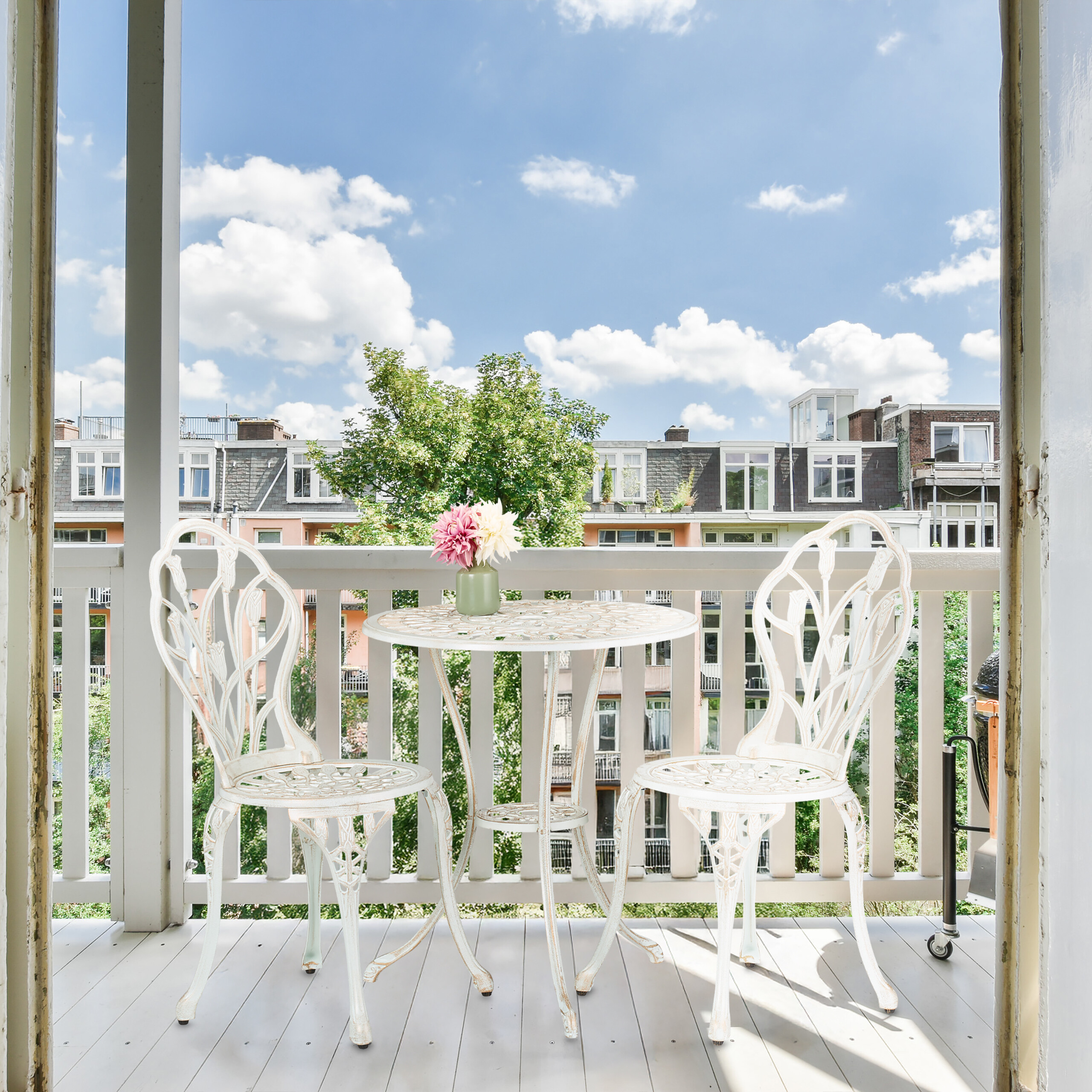 Ein weißer Balkontisch mit zwei Stühlen, Blumen in einer Vase und Blick auf Gebäude.