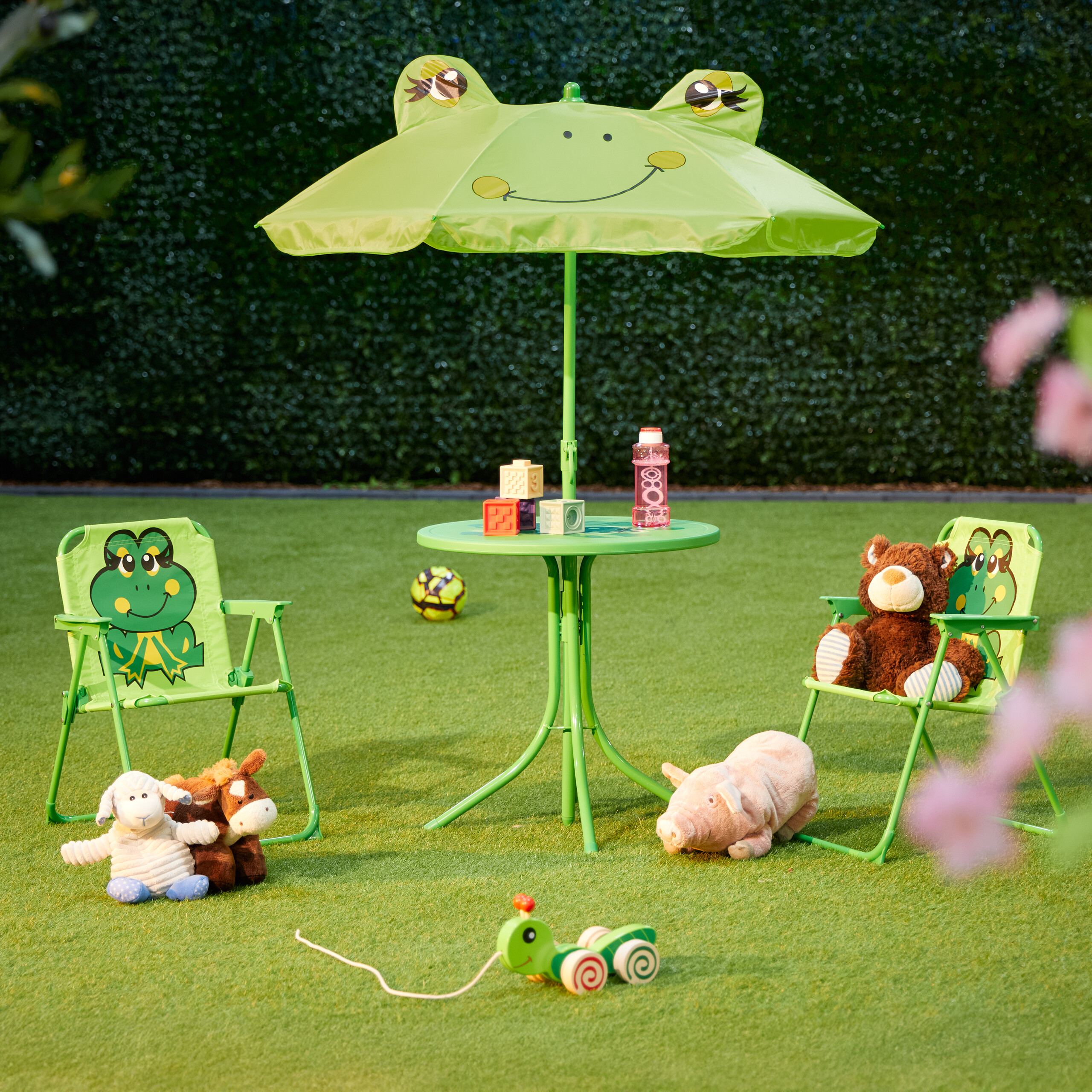 Ein grüner Frosch-Kindertisch mit Sonnenschirm und Stühlen auf grünem Rasen, umgeben von Plüschtieren.