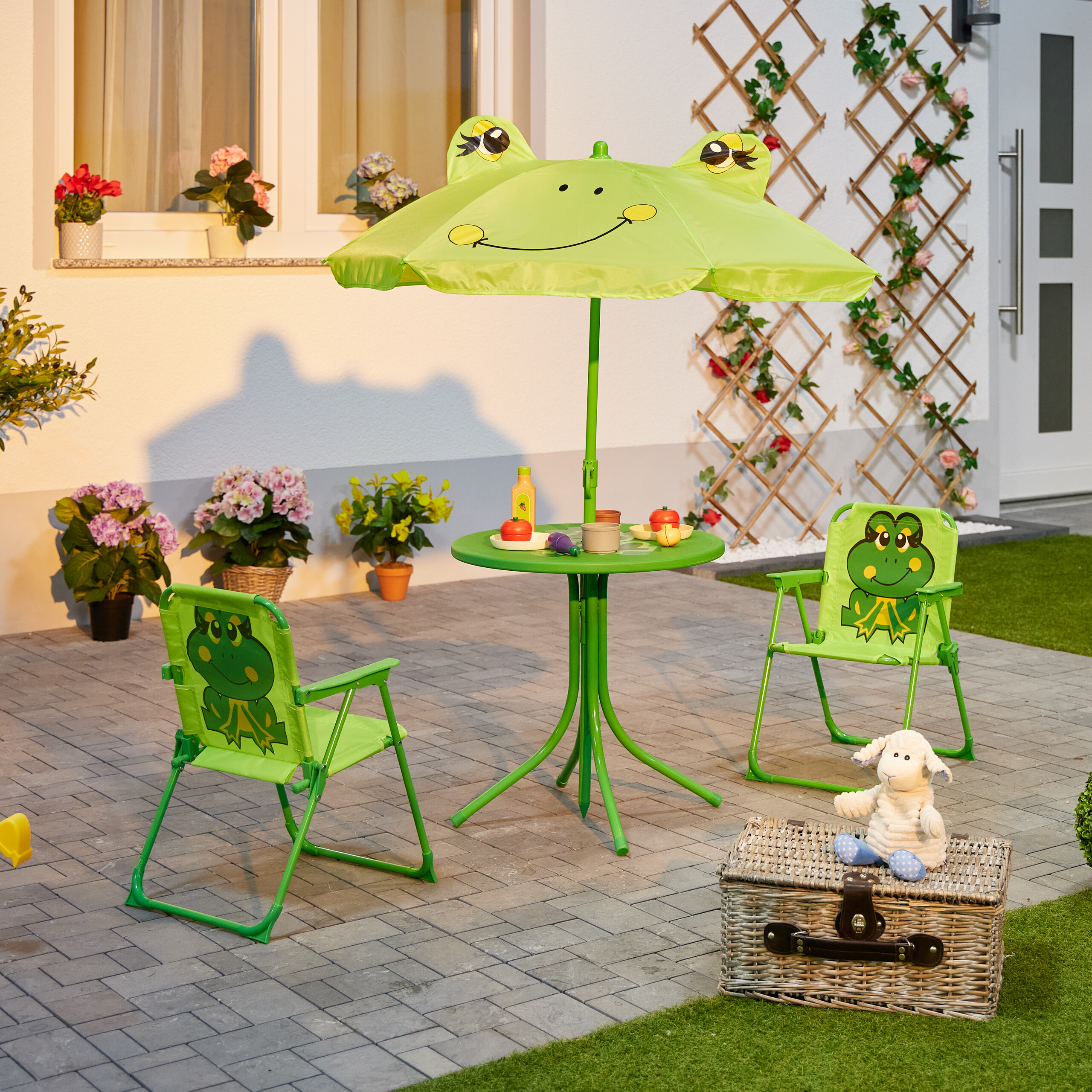 Grüner Kindersonnenschirm mit Froschgesicht, zwei Stühle, ein Tisch mit Spielzeug und ein Korb mit Stofftier.
