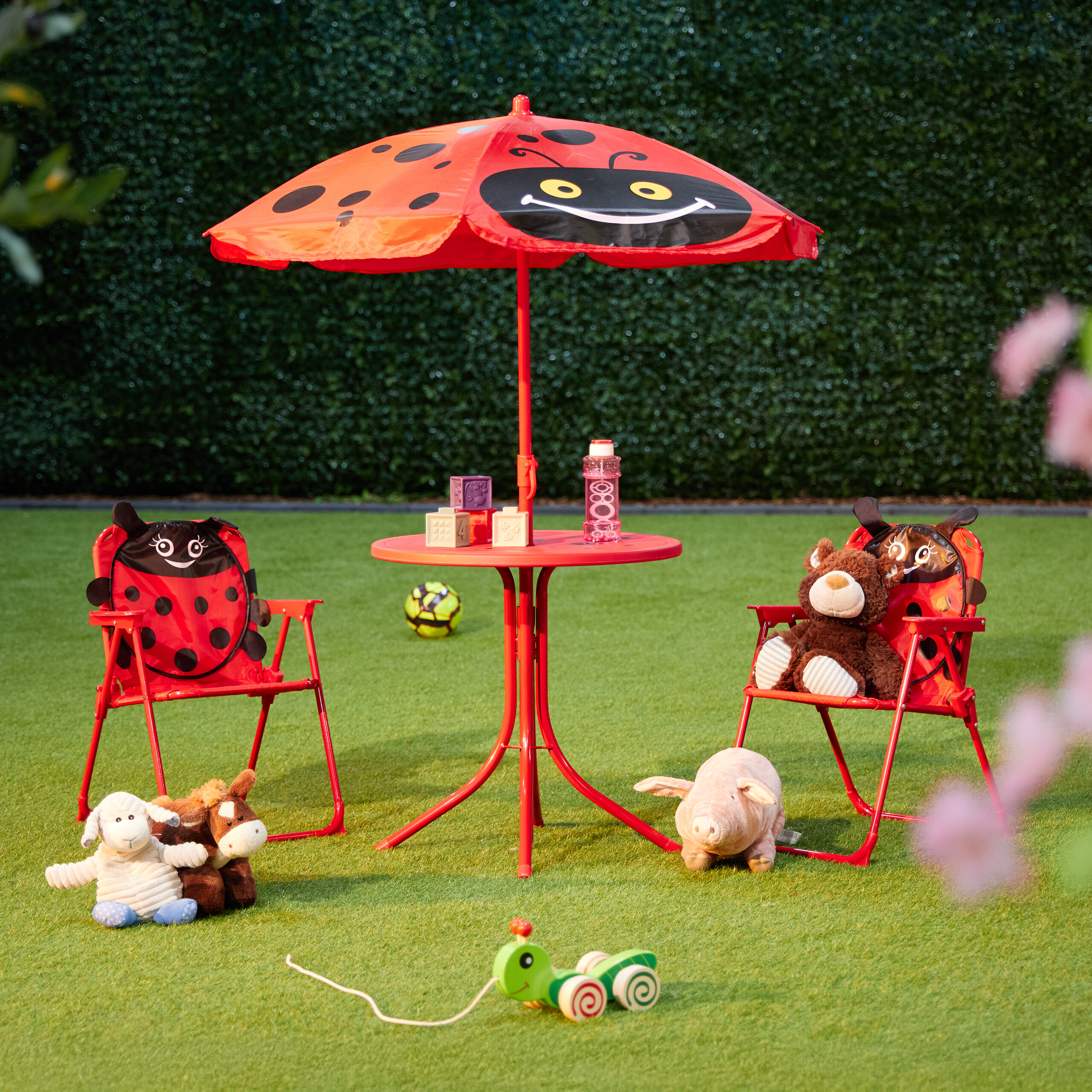 Roter Kindertisch mit Regenschirm, Stühlen und Plüschtieren auf grünem Rasen.