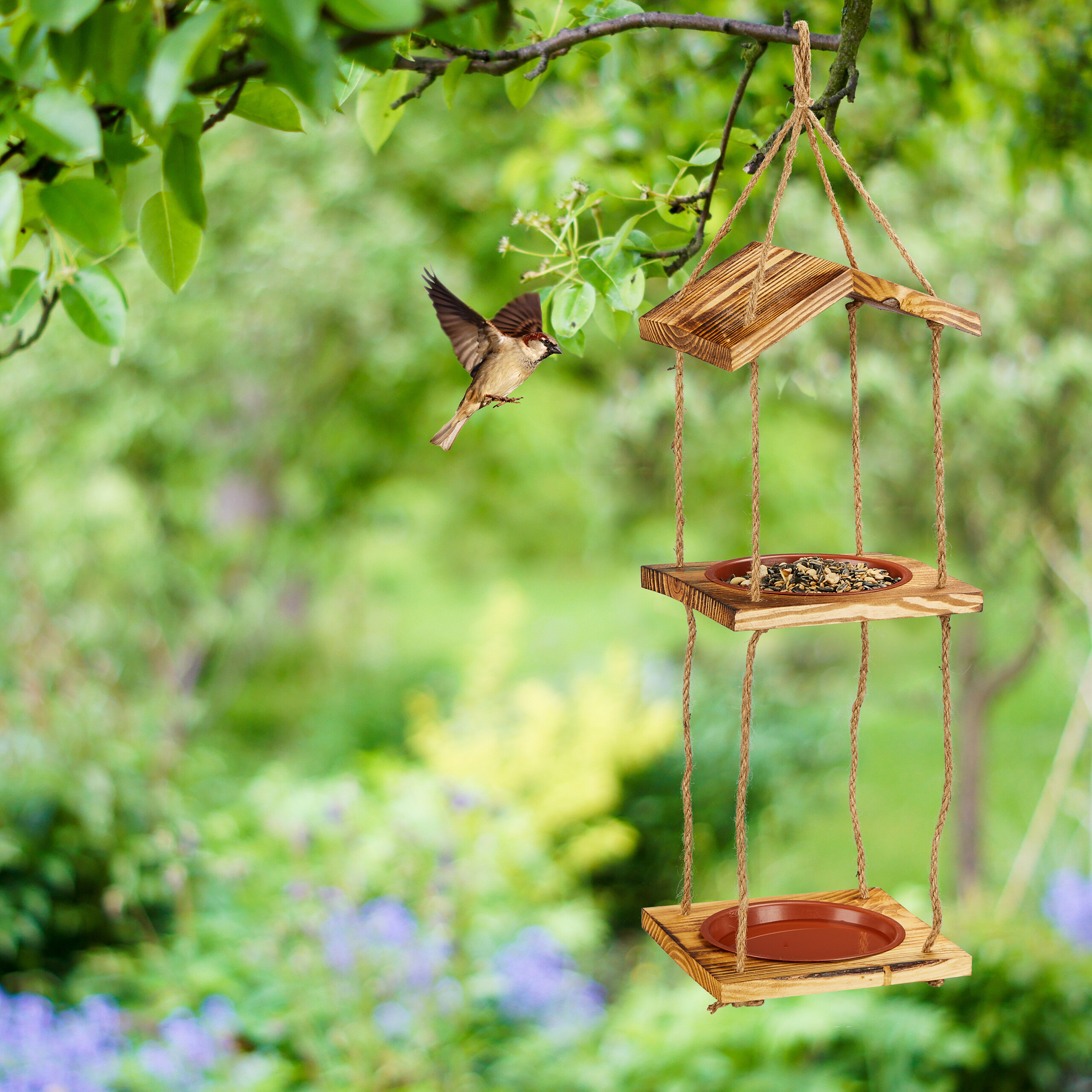 Ein kleiner Vogel fliegt zu einem hängenden Holzfutterhäuschen mit zwei Ebenen im Garten.