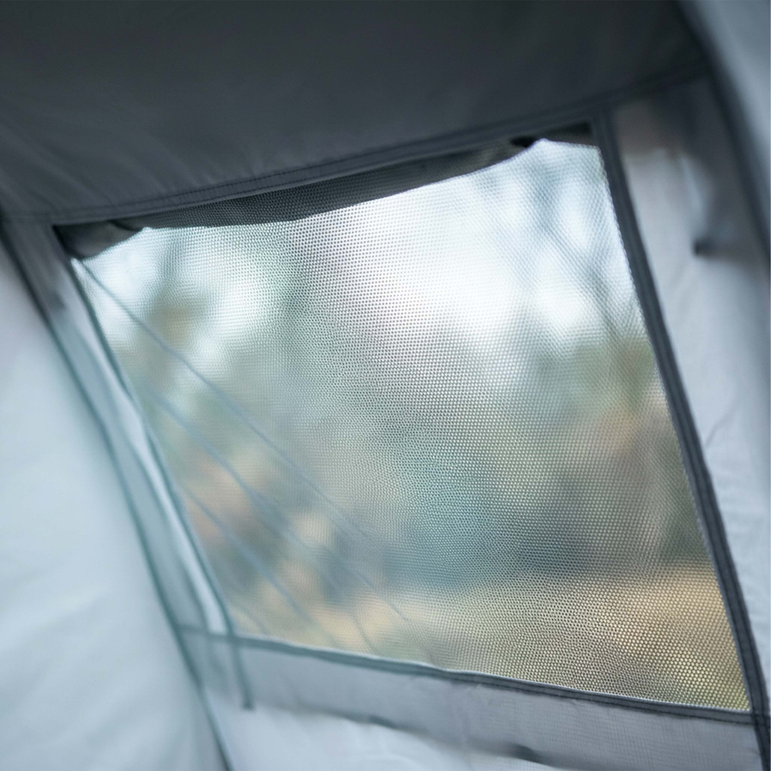 Ein Blick durch das geschlossene, graue Netzfenster eines Zeltes, wobei die Natur draußen verschwommen ist.
