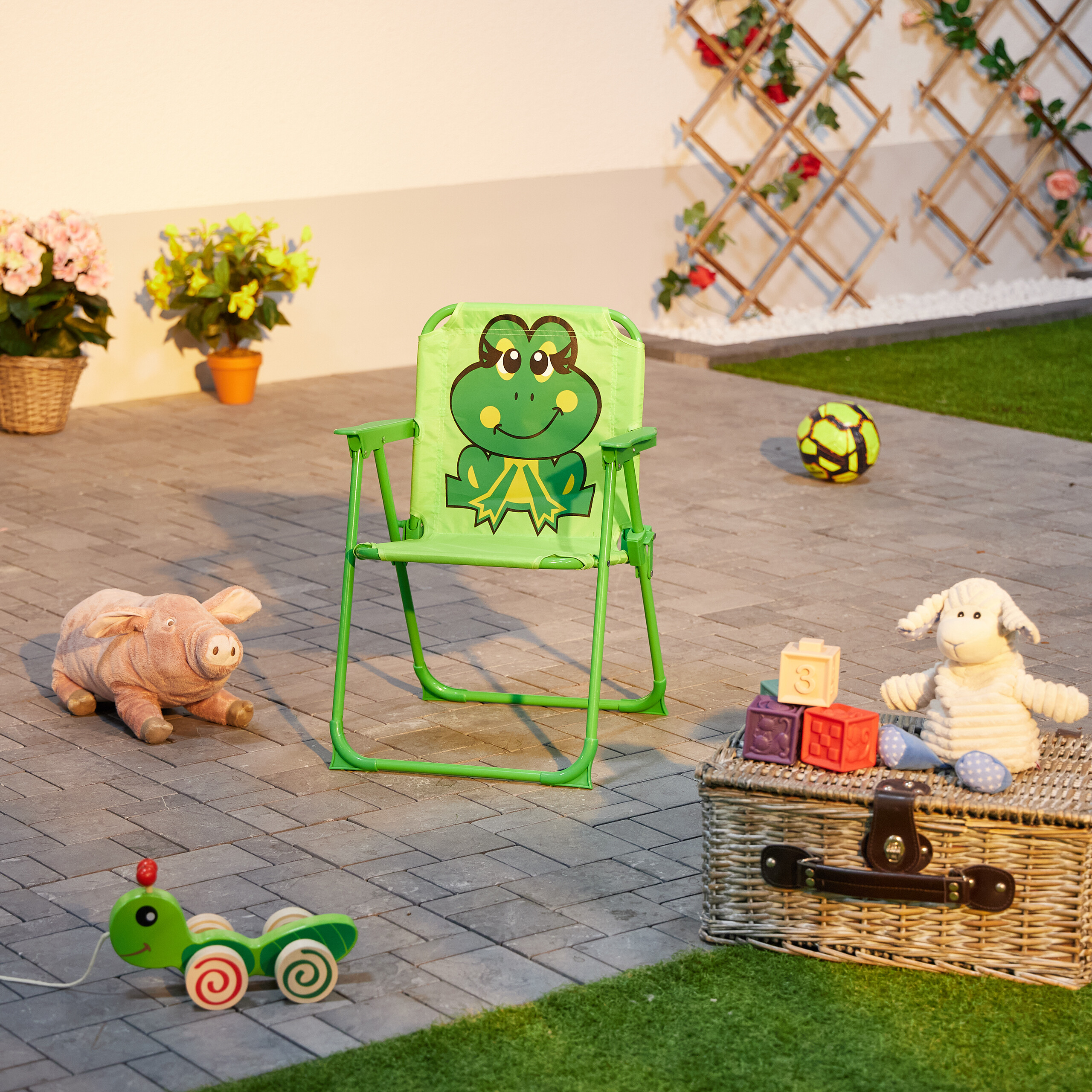 Grüner Frosch-Kinderstuhl auf gepflasterter Terrasse mit Spielzeug, Korb und Blumen im Hintergrund.