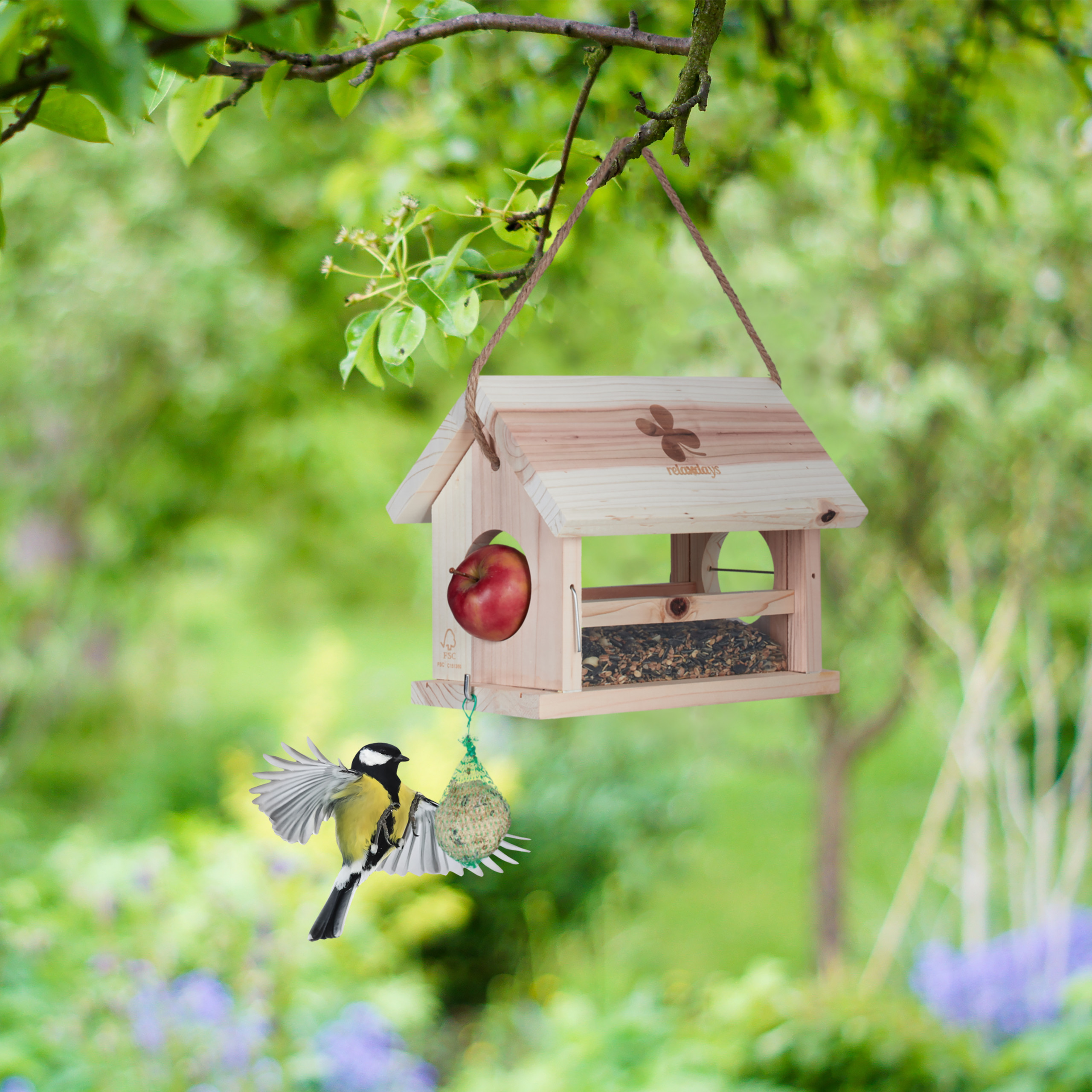 Eine Meise fliegt zu einem hölzernen Vogelhaus mit Äpfeln und Futter im grünen Garten.