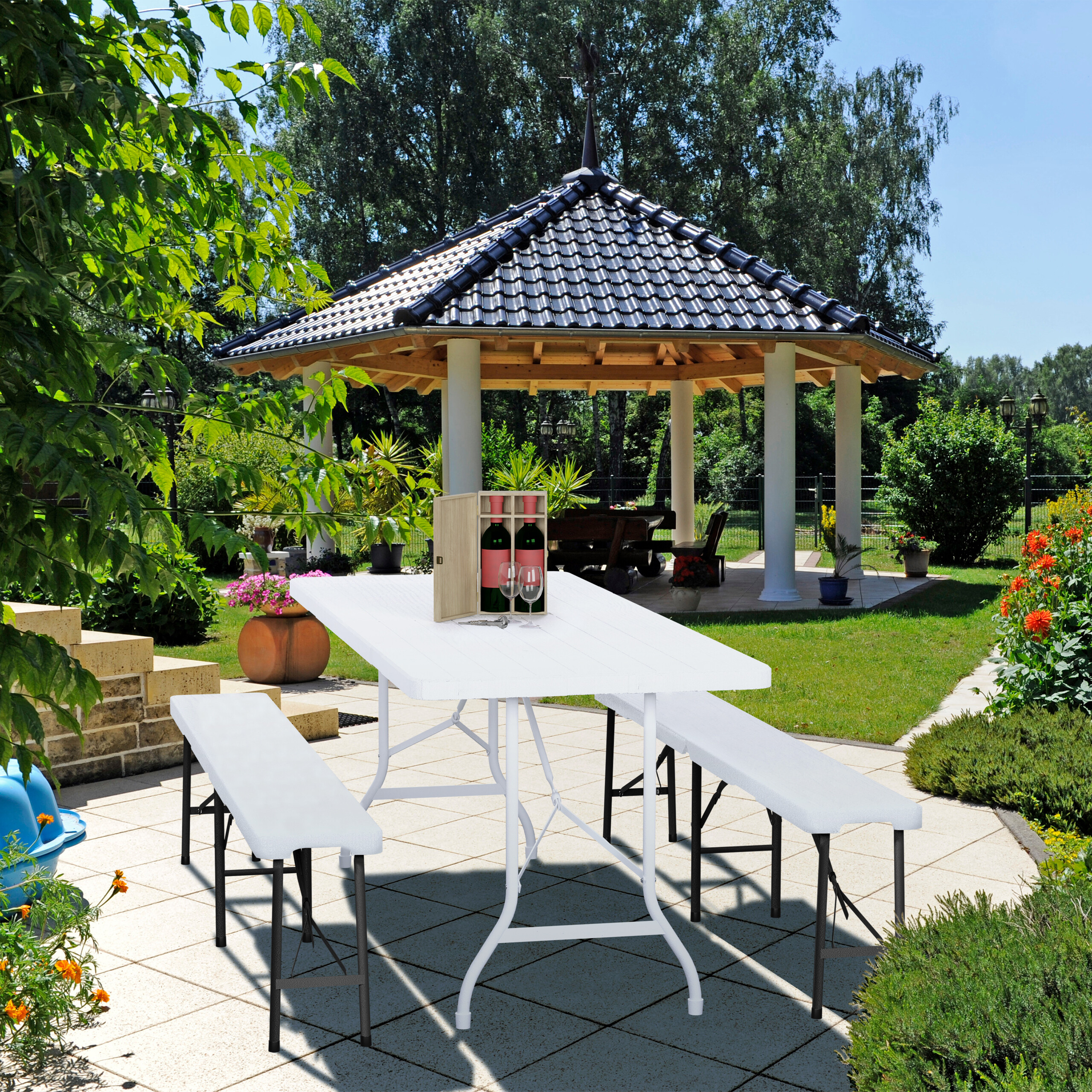 Ein weißer Klapptisch und zwei Bänke auf einer Terrasse mit Weinkiste und Gläsern im grünen Garten.